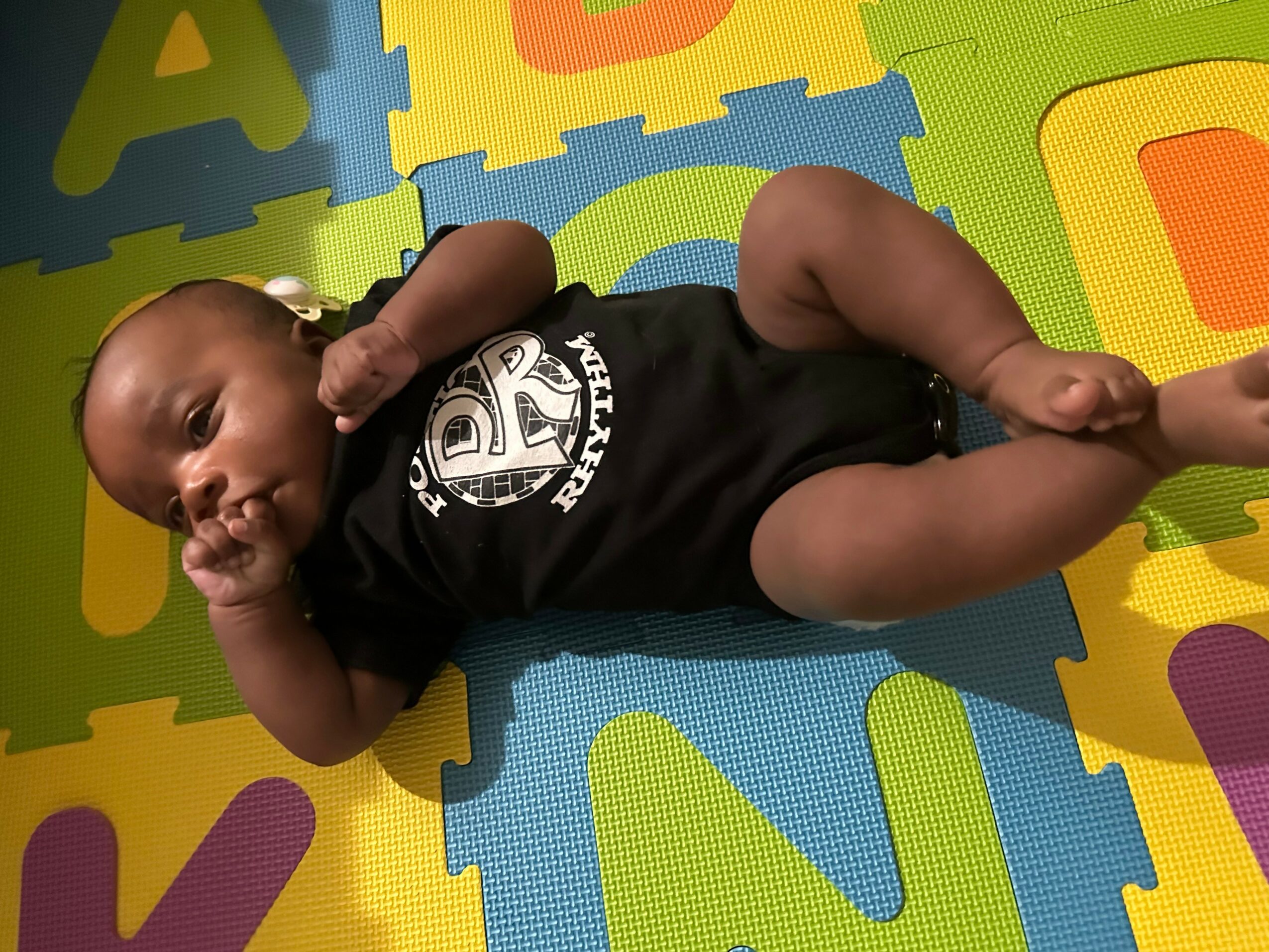 Baby lying on colorful foam mat, wearing a black onesie, looking to the side, with one hand near ear.