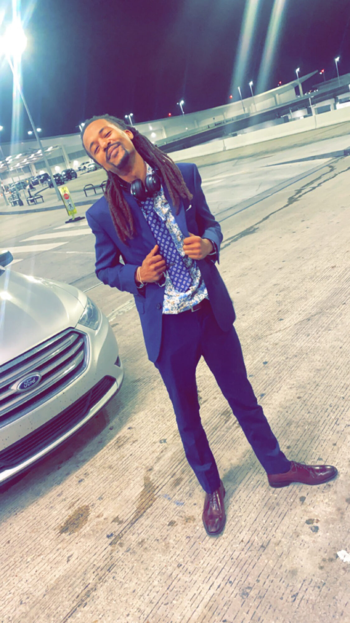 Person with long hair and glasses smiling, standing outdoors at night, wearing a blue blazer and patterned shirt, near a car.