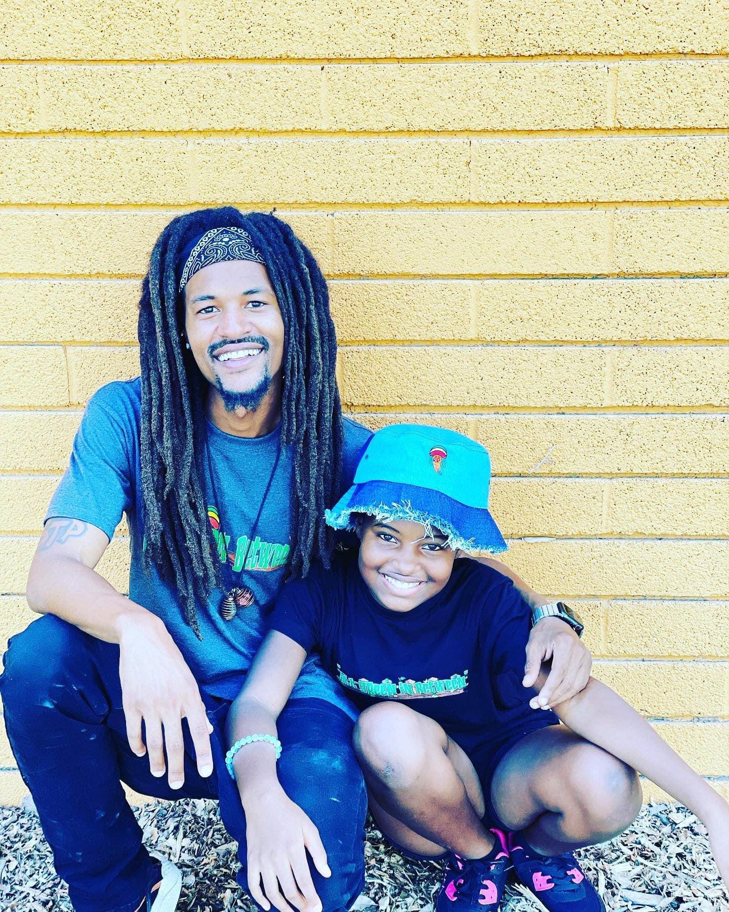 Two smiling people, one adult and one child, sitting against a yellow wall, with the adult's arm around the child.