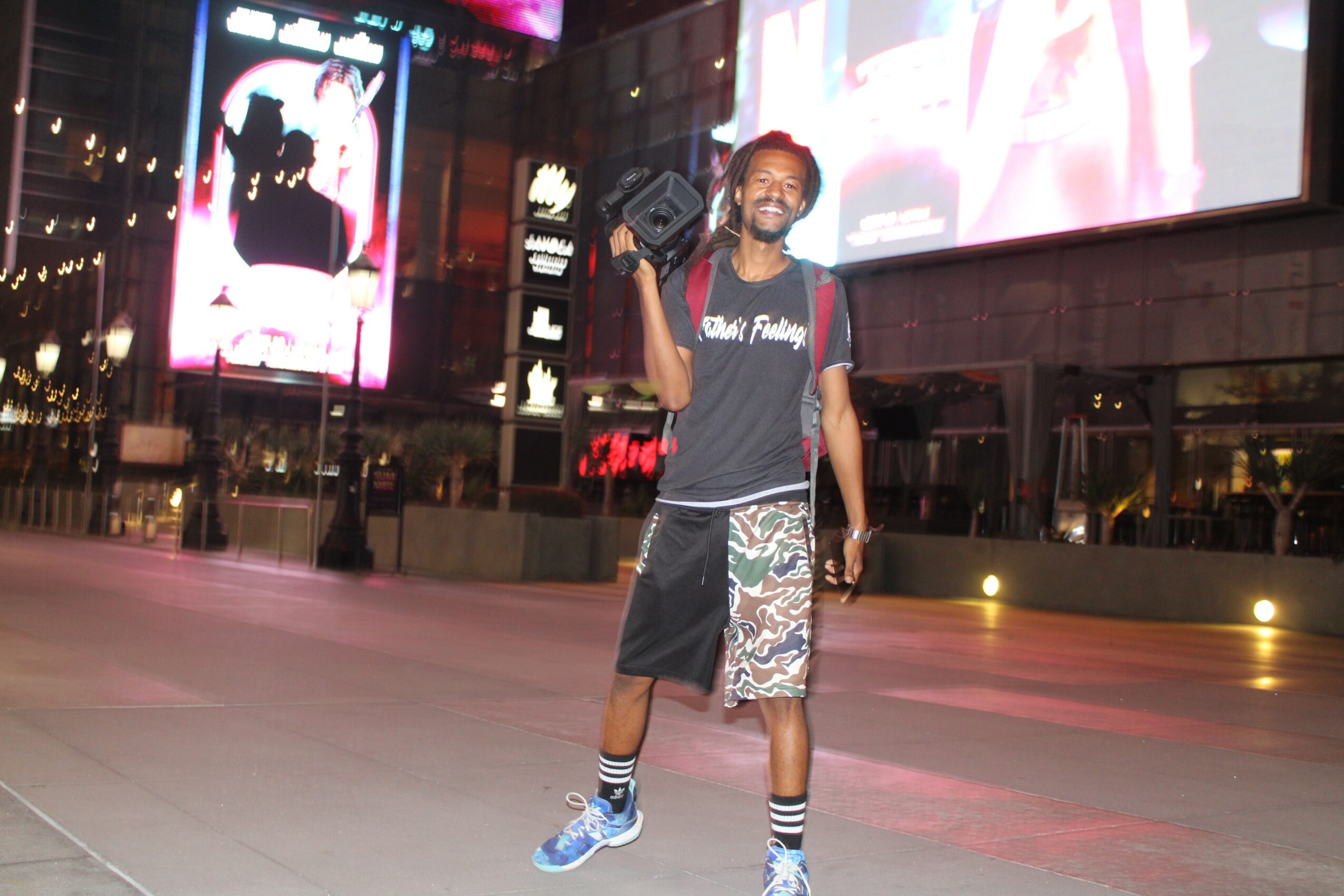 Young man standing outdoors at night, smiling, carrying a backpack, in an urban area with bright billboards and buildings behind him.