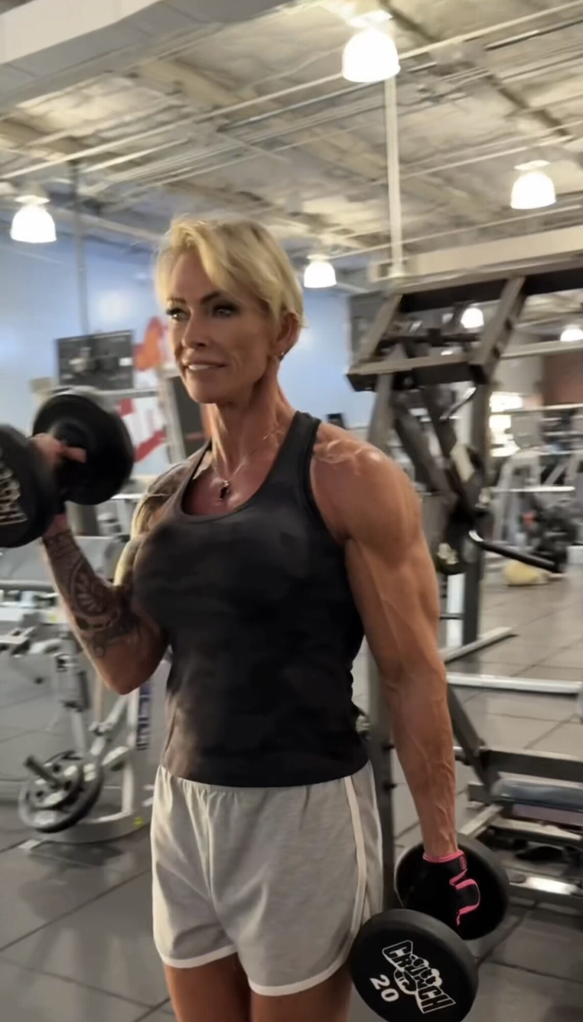 Woman lifting dumbbell in gym, wearing black tank top and white shorts, with gym equipment in background.