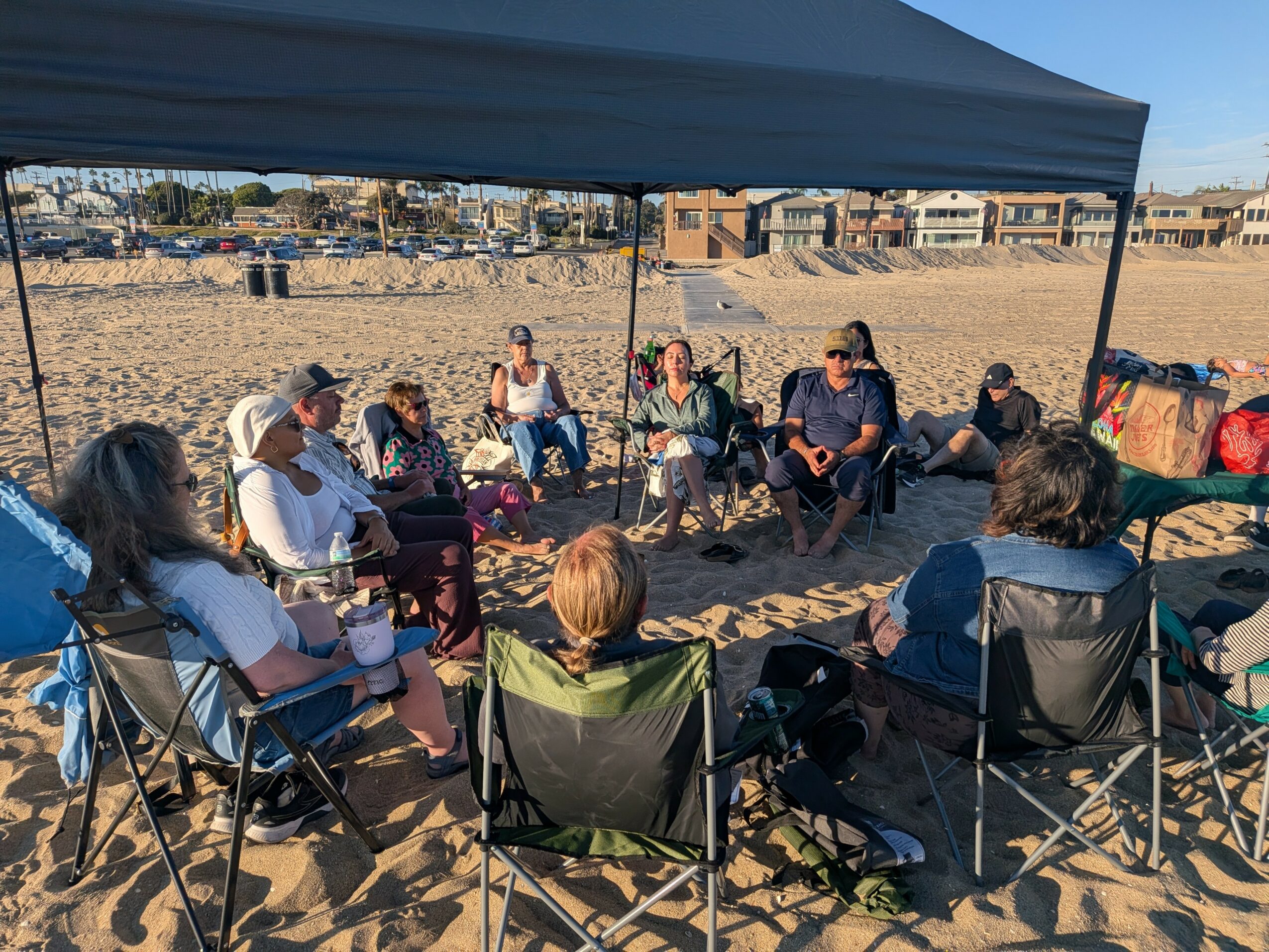 Group of people sitting in a circle under a canopy on a sandy beach, with buildings and a clear sky in the background.