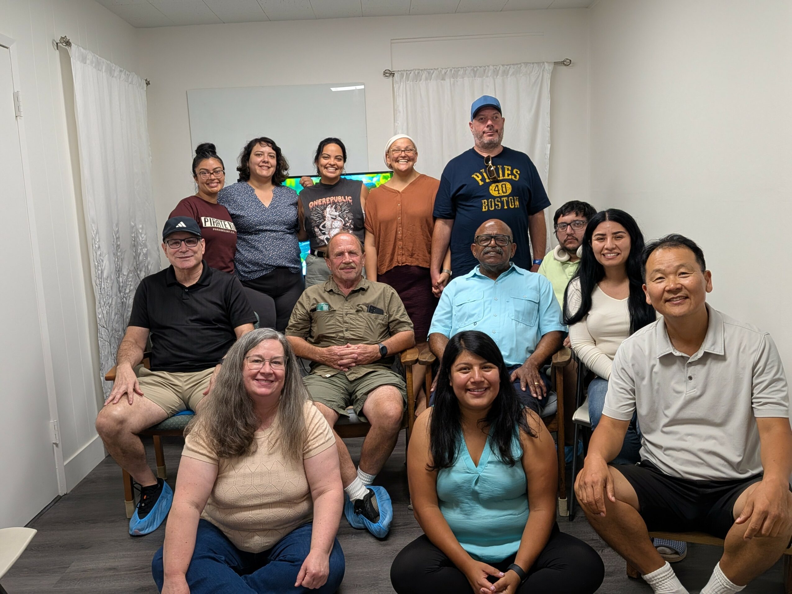 Group of twelve people posing indoors, some sitting and some standing, in a room with white walls and curtains.