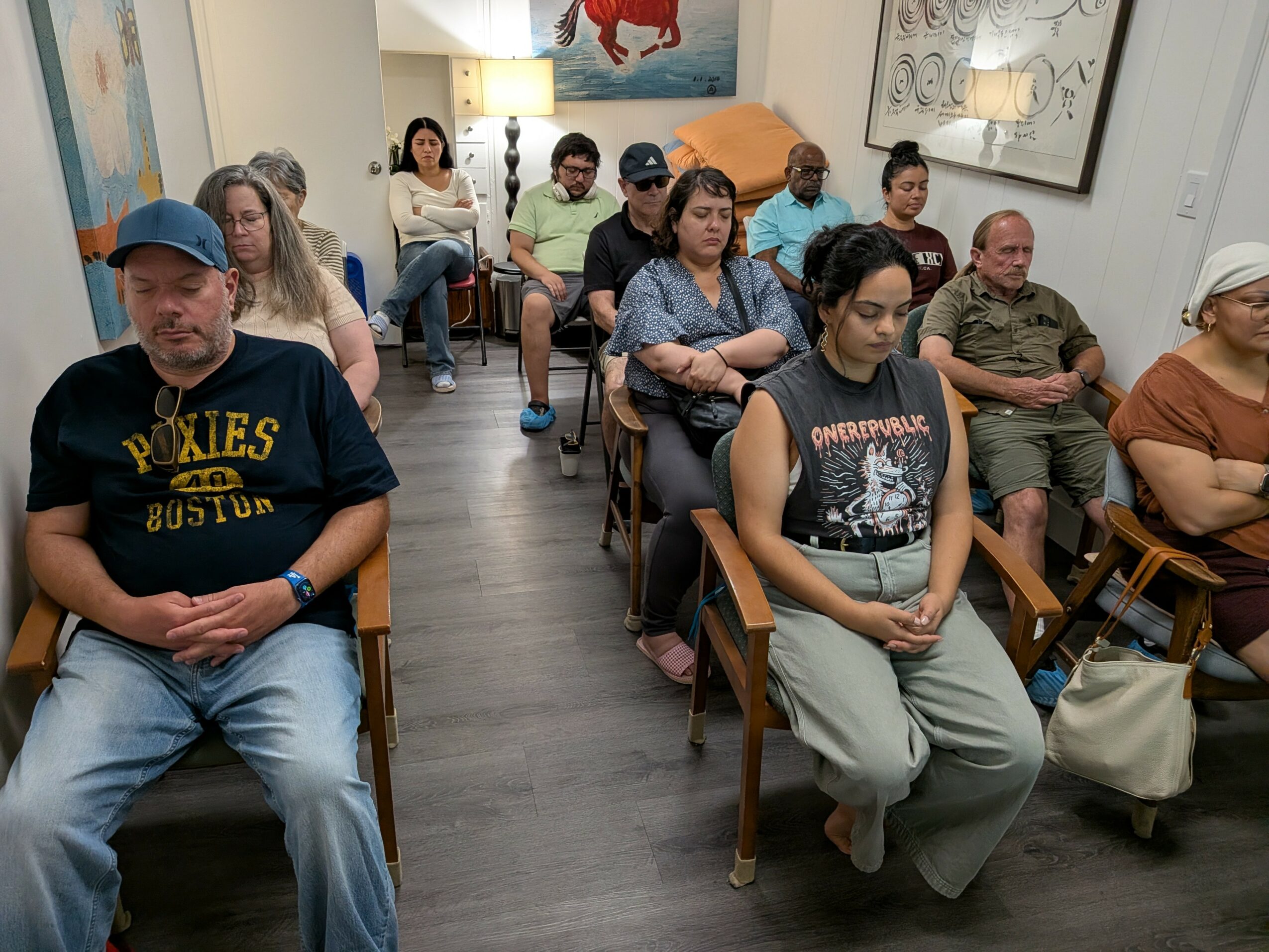 Group of people seated in a room, some with eyes closed, listening attentively, with artwork on the walls.