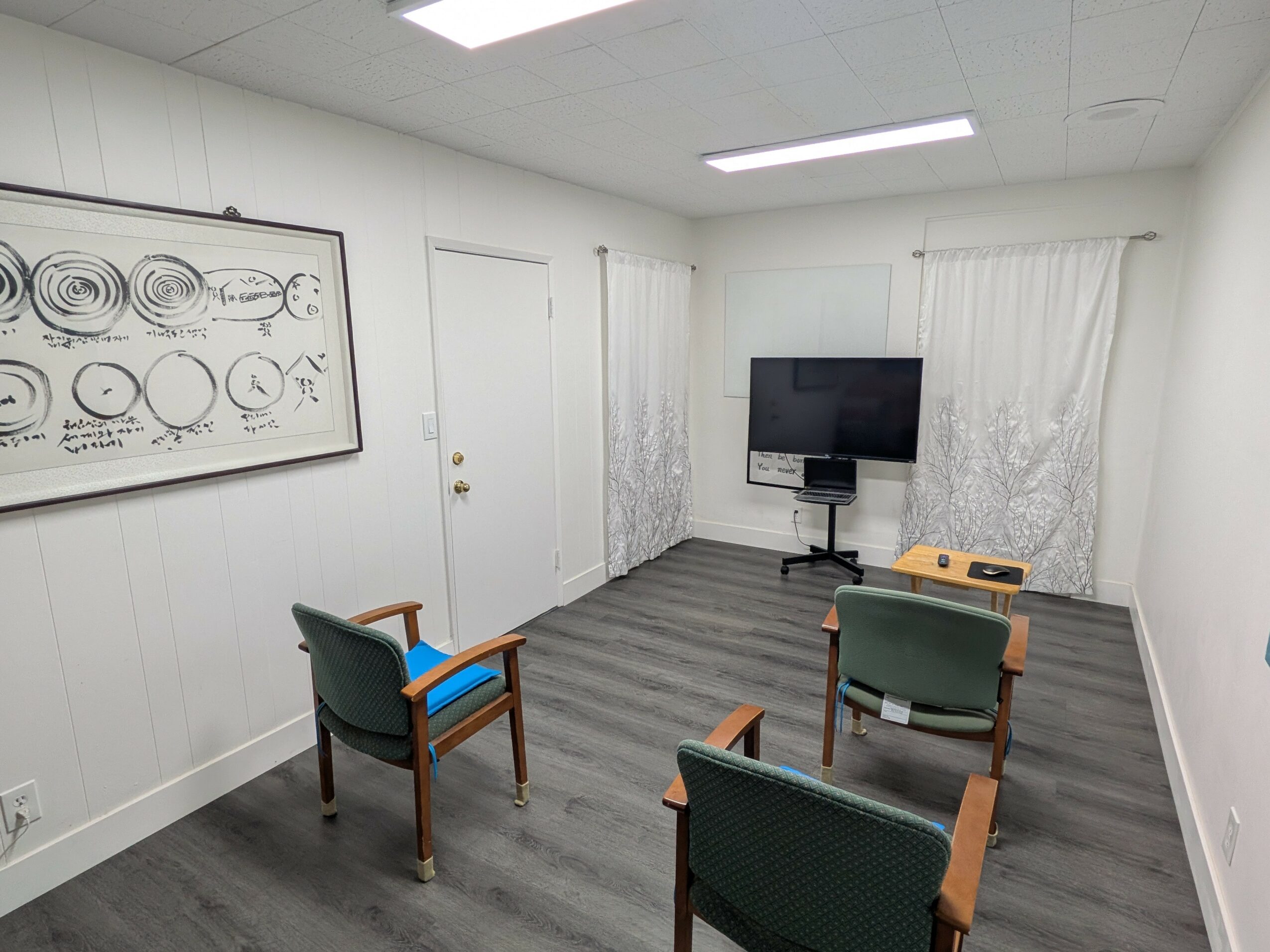 Empty room with four chairs facing a wall-mounted TV, white walls, gray flooring, and a framed diagram on the wall.
