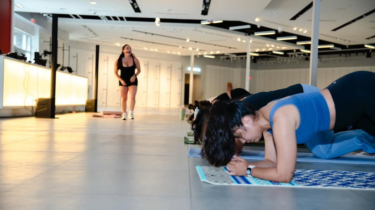 Person doing a plank exercise on a mat in a spacious indoor gym, with a woman walking in the background.