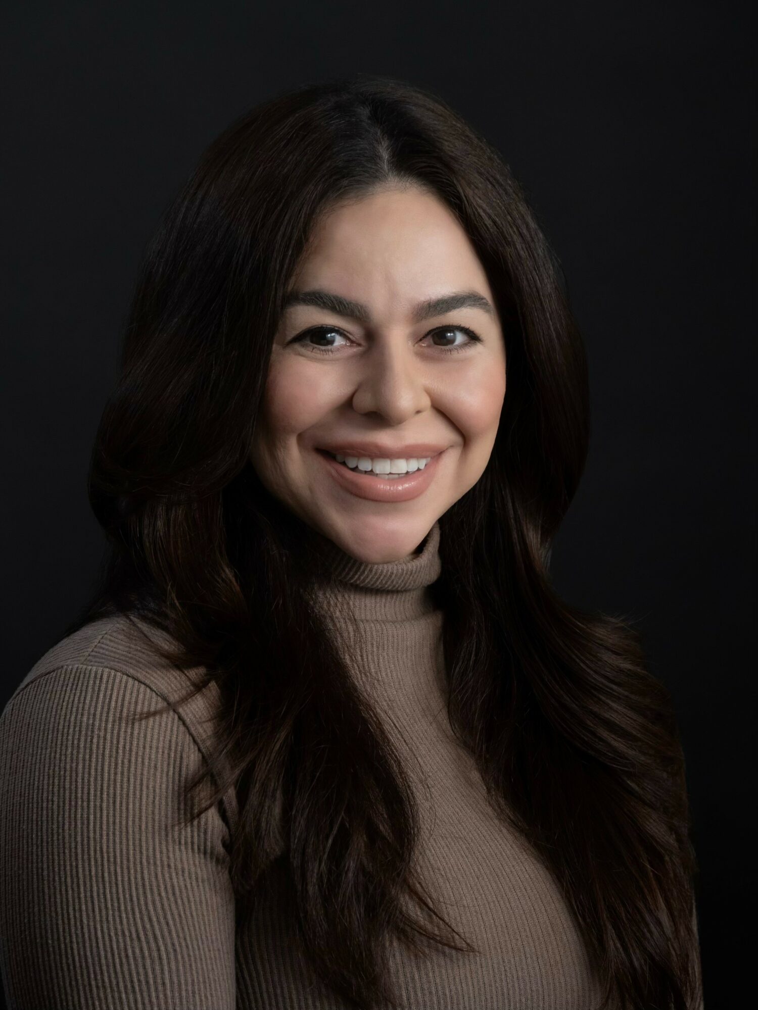 Portrait of a smiling woman with long dark hair wearing a beige turtleneck against a dark background.