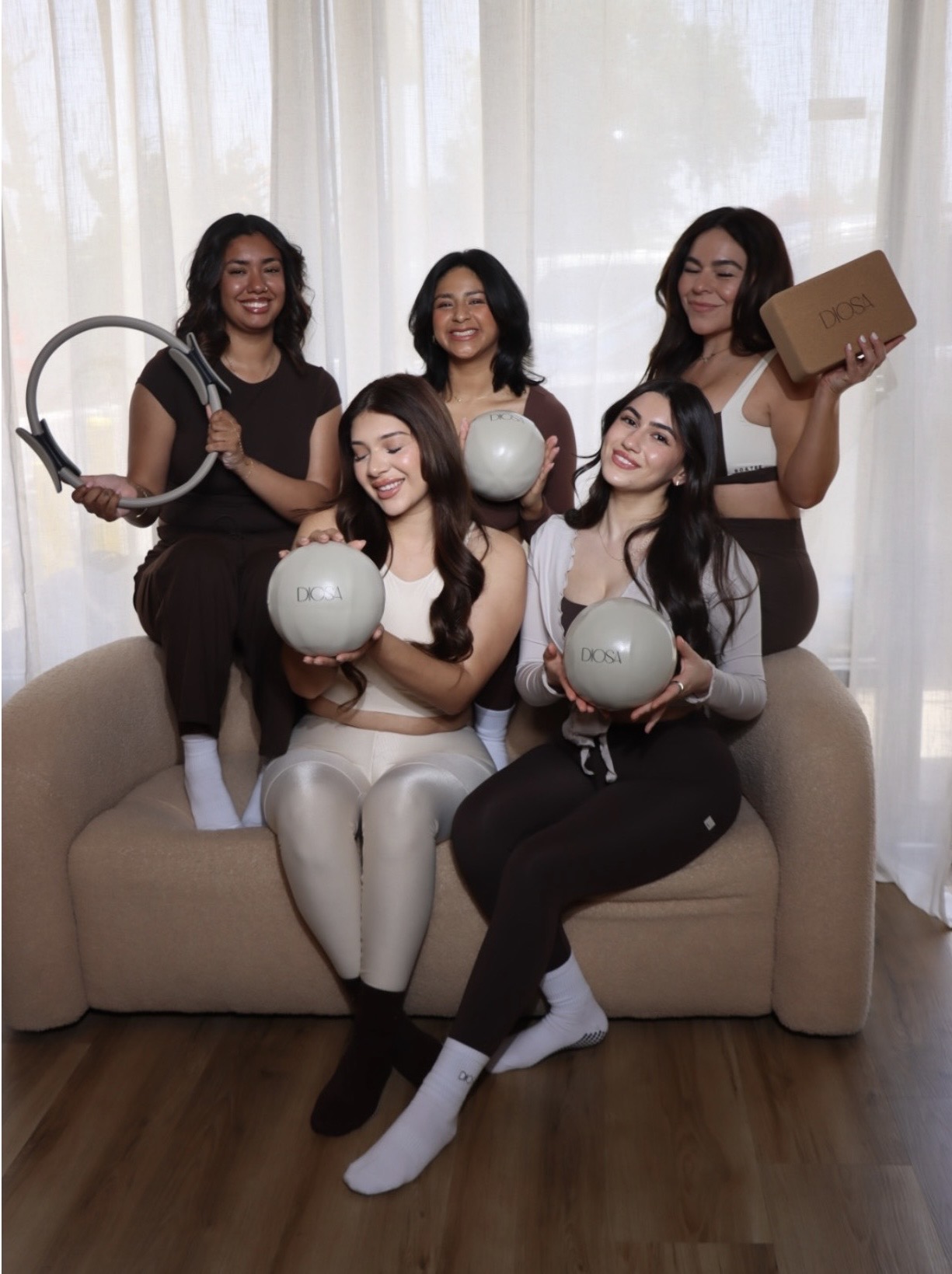 Five women sitting and standing on a sofa, holding spherical objects and a cardboard box, smiling indoors with curtains behind them.