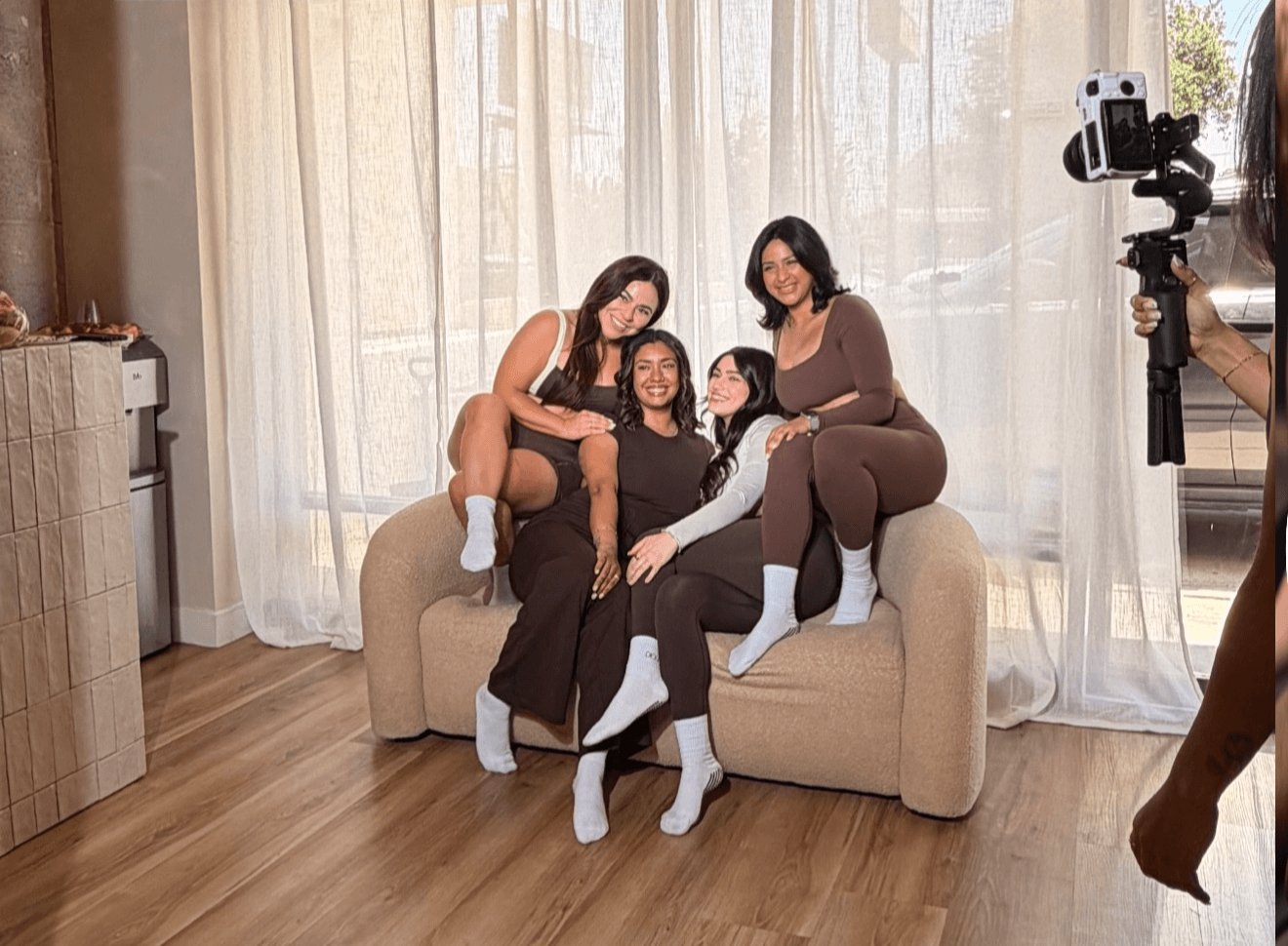 Three women sitting on a beige sofa in a room with large windows and sheer curtains, smiling at the camera.