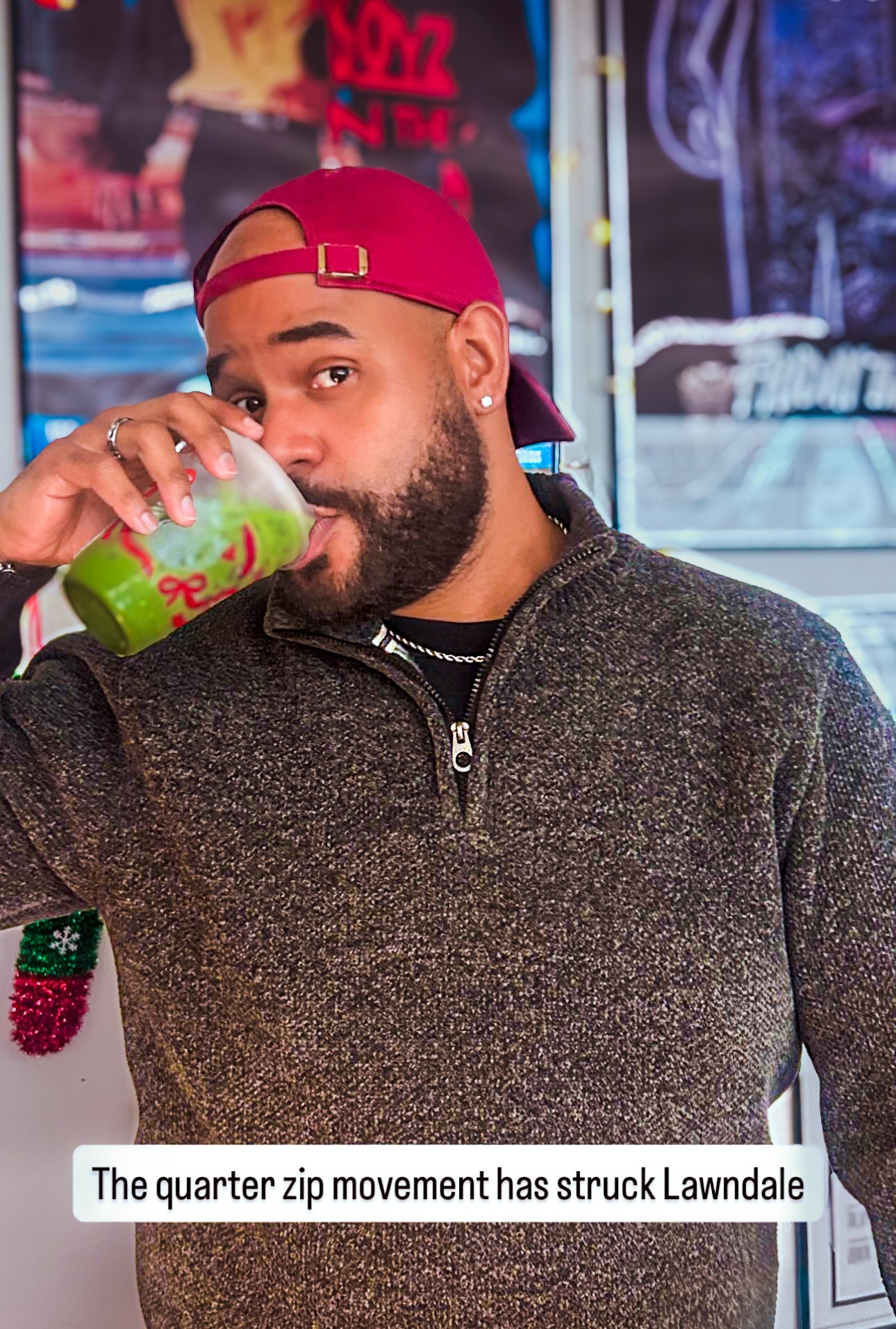 Man with beard wearing a red cap and dark sweater drinks from a green cup in a colorful setting.