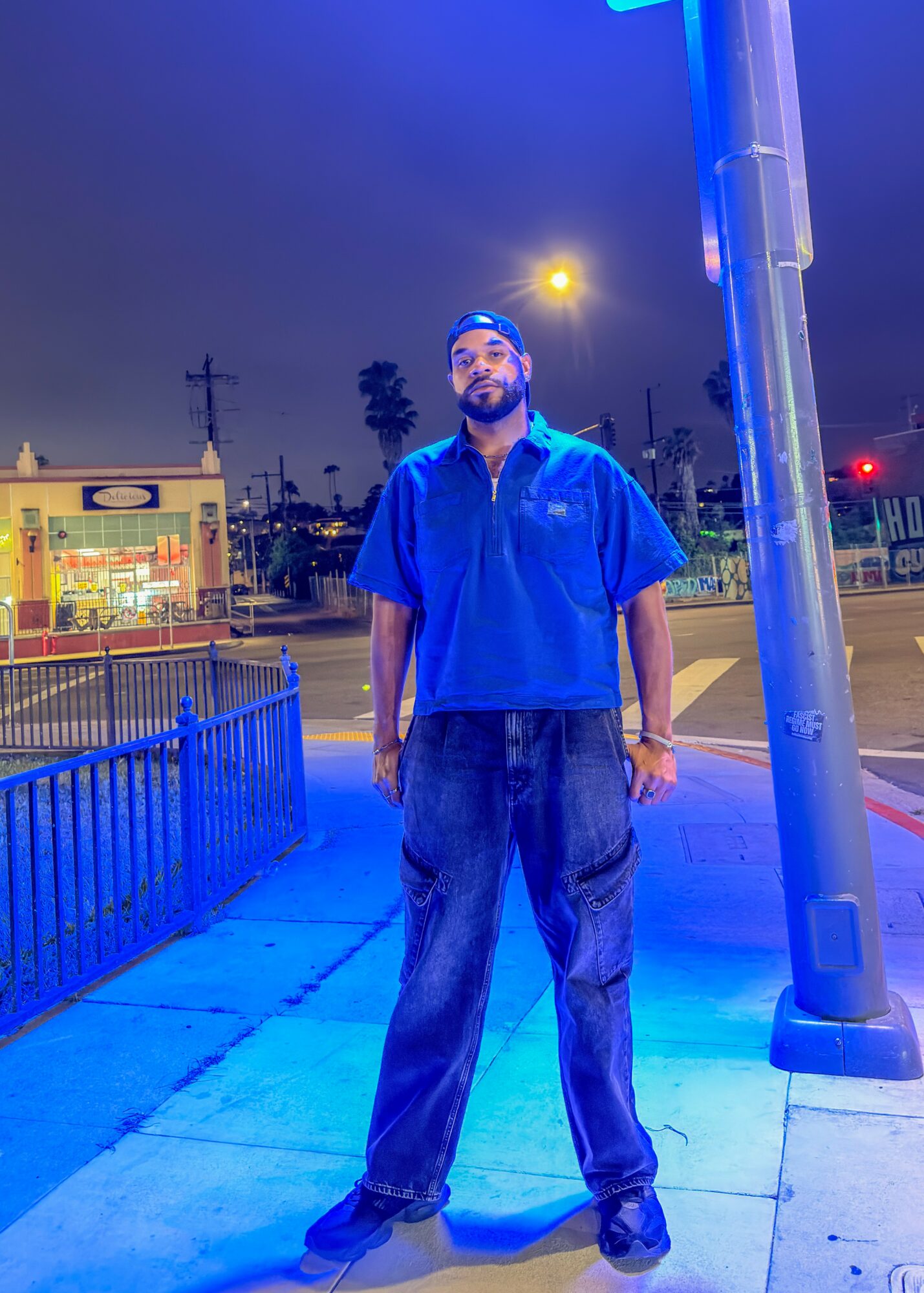 Man standing on sidewalk at night, illuminated by blue streetlight, with buildings and street in background.