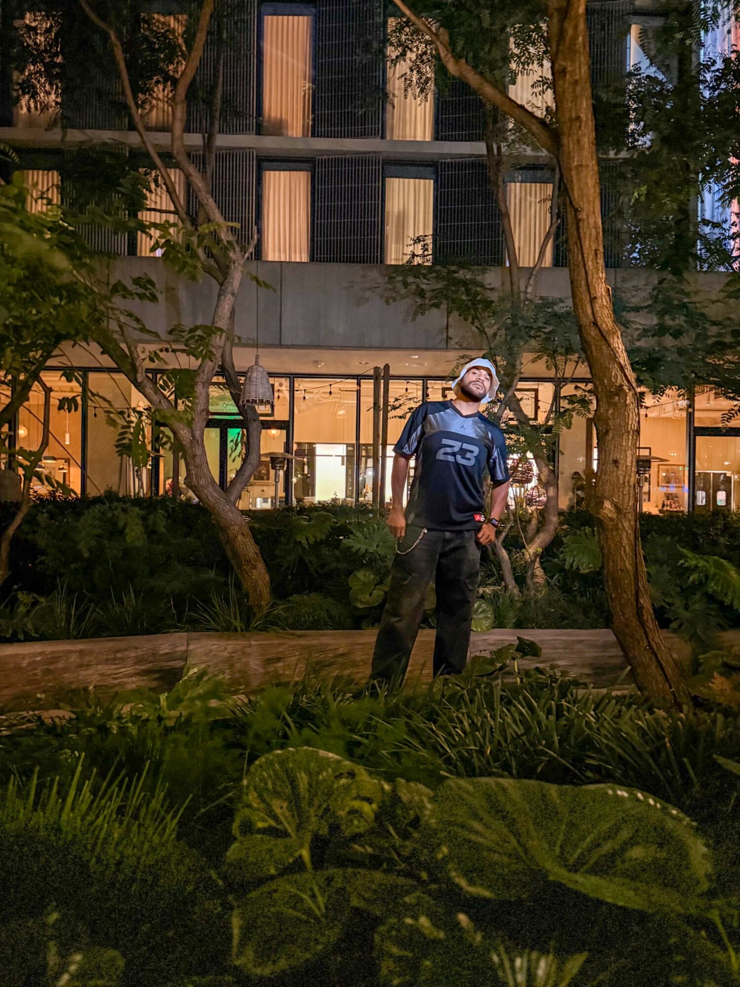 Person wearing a mask and sports jersey standing outdoors near trees and plants at night.
