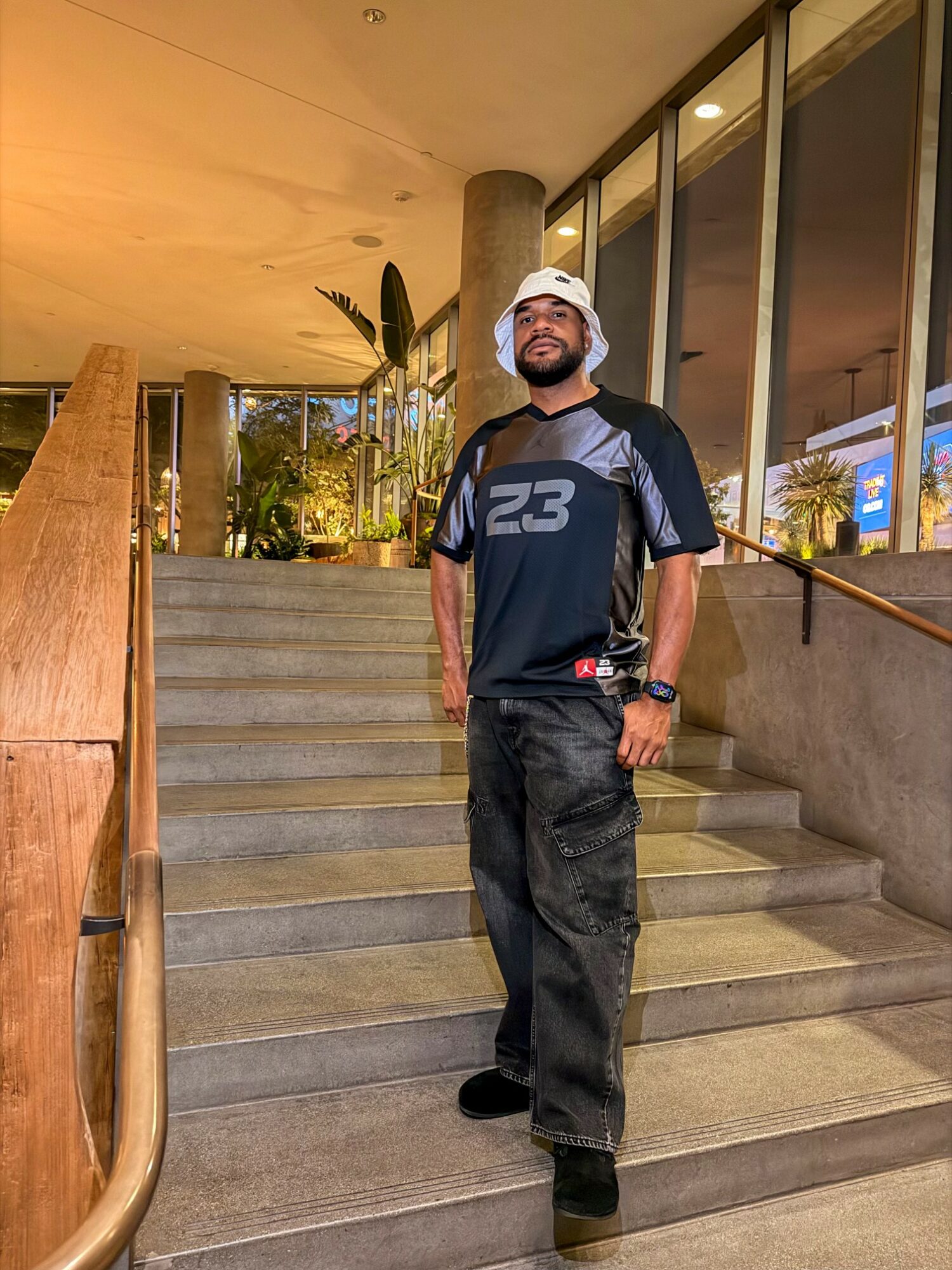 Man standing on stairs indoors, wearing a white cap, black pants, and a black and blue shirt, with glass windows behind him.