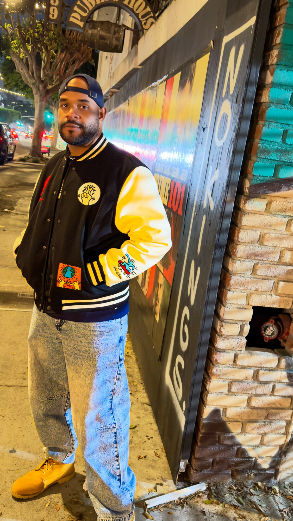 Person standing outdoors at night, wearing a black and yellow jacket, jeans, and yellow shoes, near a colorful wall.