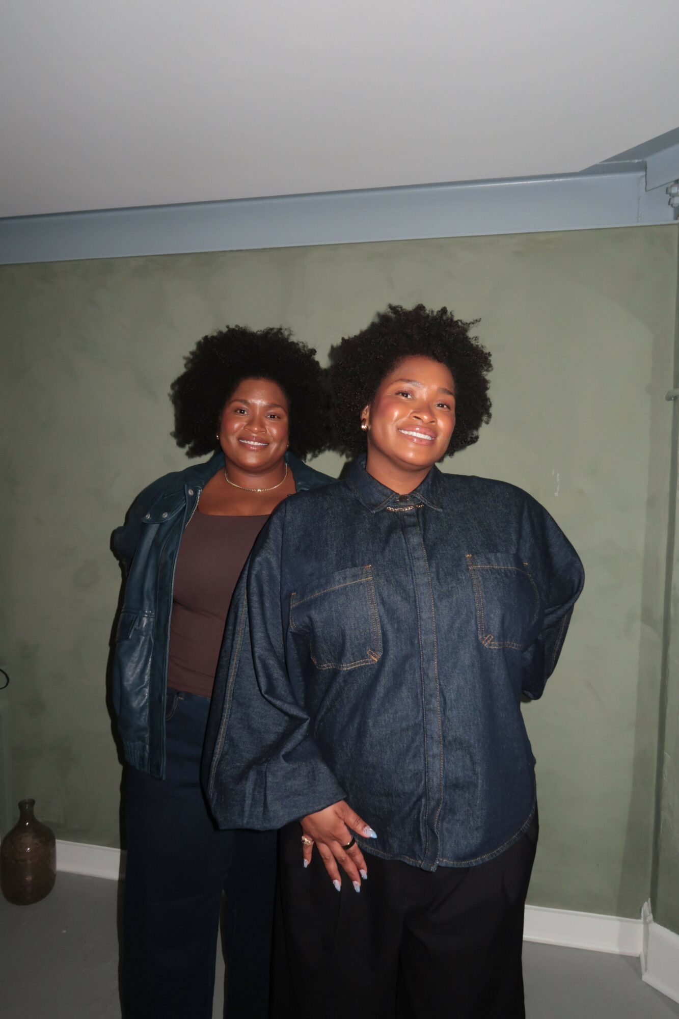 Two women standing indoors, one with curly hair wearing a denim jacket, the other with short hair in a dark top, smiling.