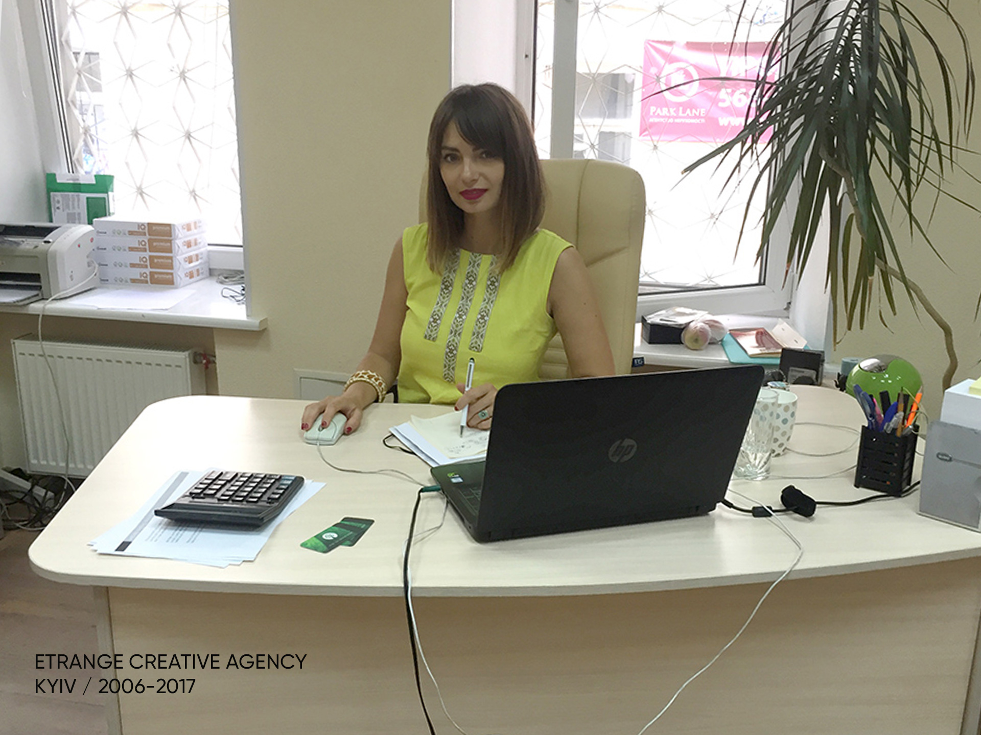 Woman sitting at a desk with a laptop, papers, and office supplies, in a bright office with a window and plant.