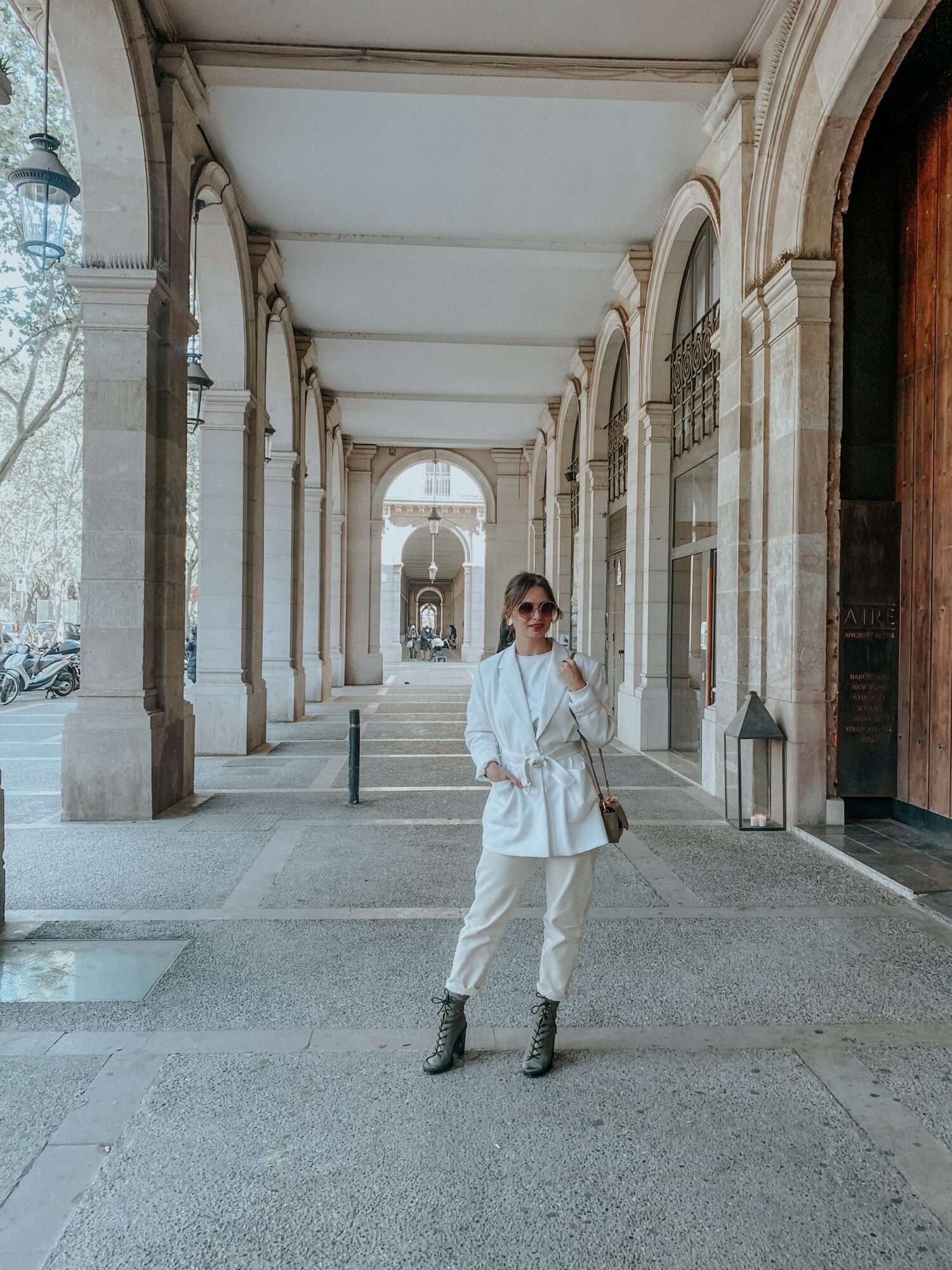 Person standing in a covered walkway with arches, wearing white outfit and sunglasses, holding a bag, with columns and trees in background.