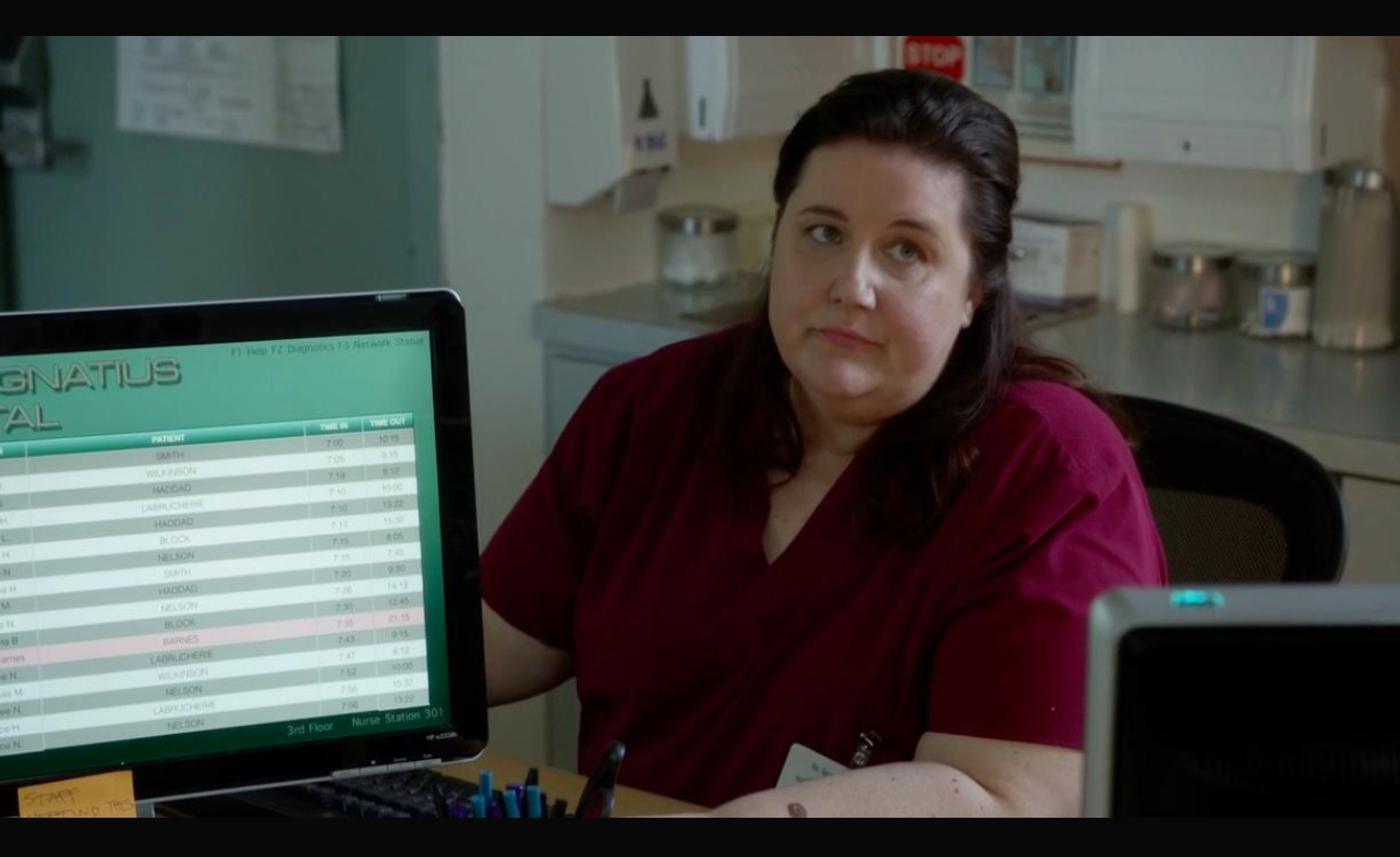 Woman with dark hair in a maroon top sitting at a desk with a computer monitor in a medical or office setting.