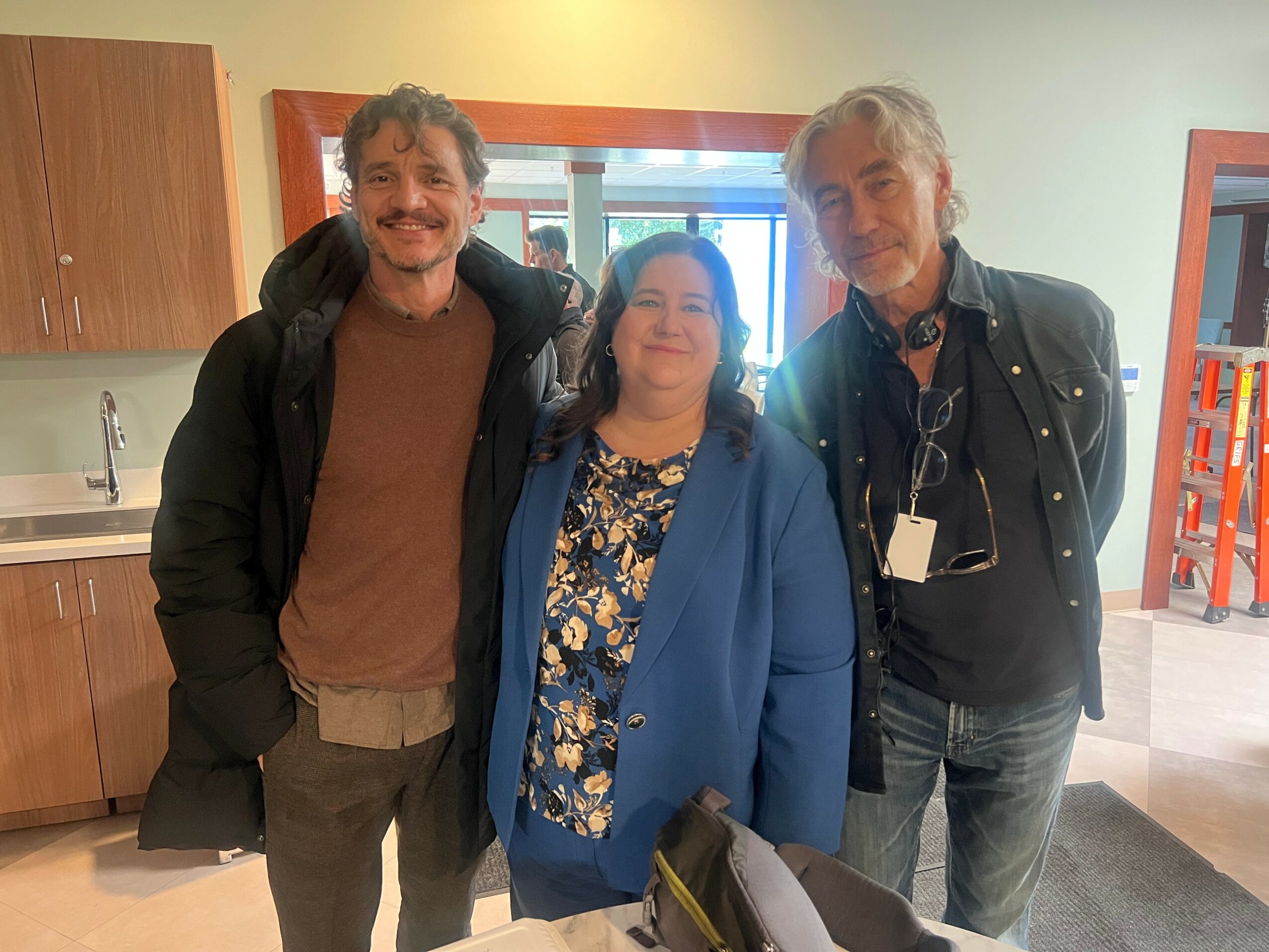 Three people standing indoors, smiling, with a kitchen and window in the background.