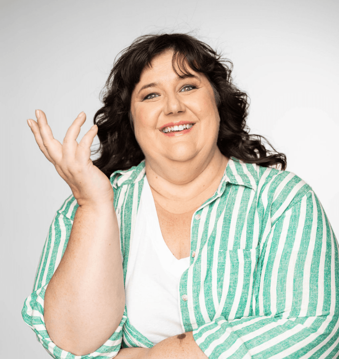 Smiling woman with dark curly hair wearing a green and white striped shirt and white top, gesturing with her hand.