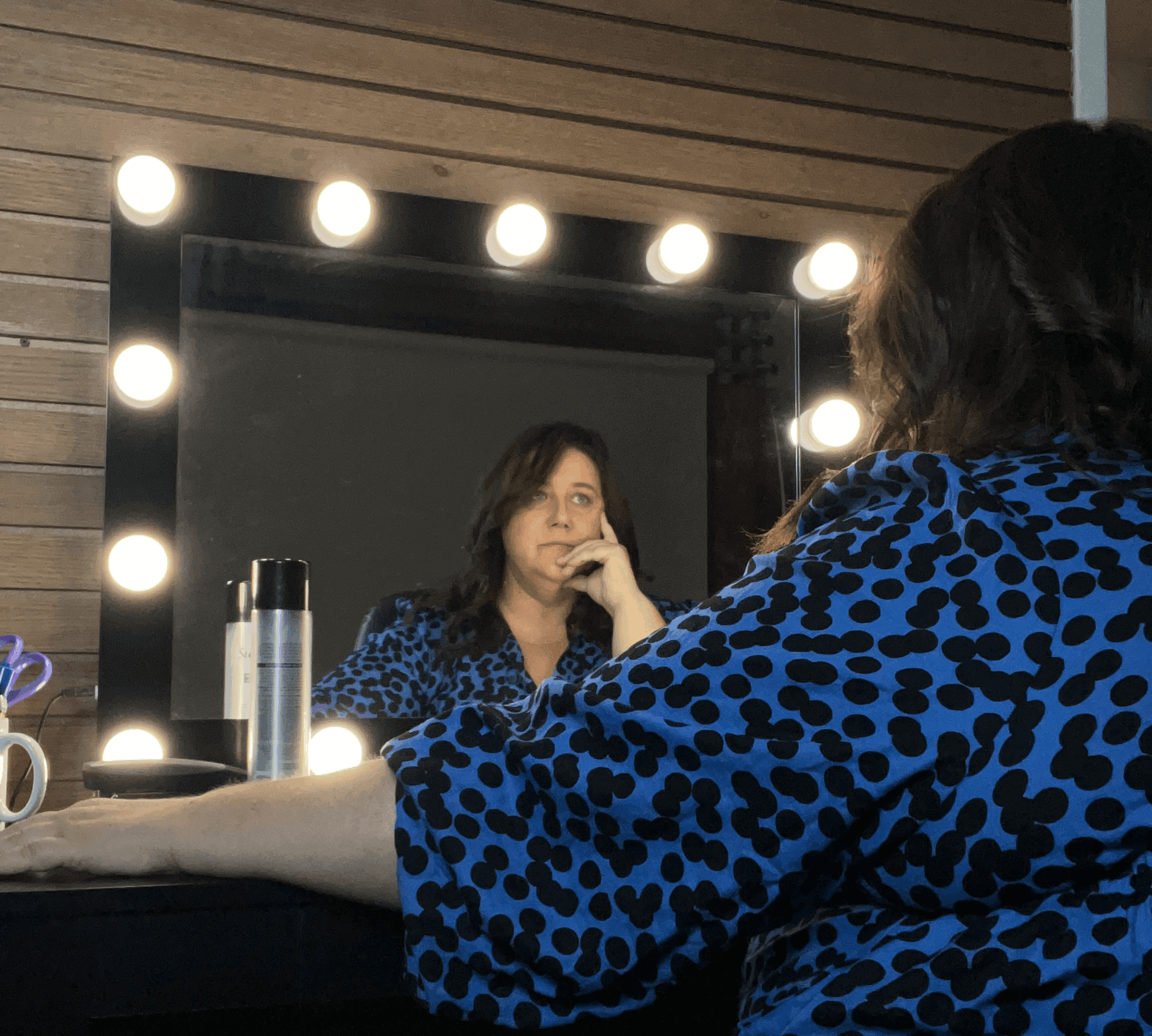 Person with dark hair and blue patterned top looking at mirror with lights, sitting at a vanity table.