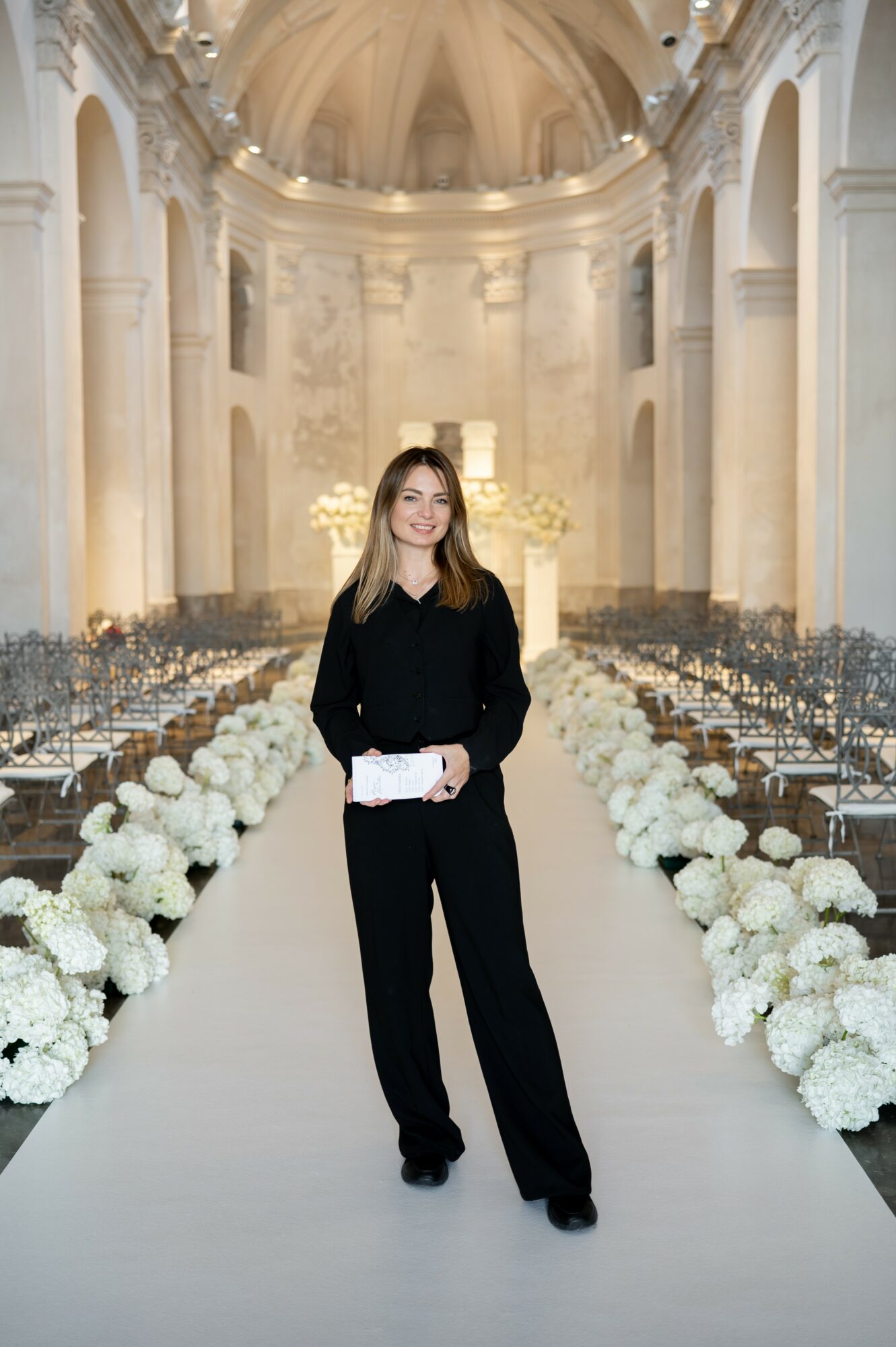 Woman in black standing in a decorated church aisle with white flowers, ornate architecture, and a high ceiling.