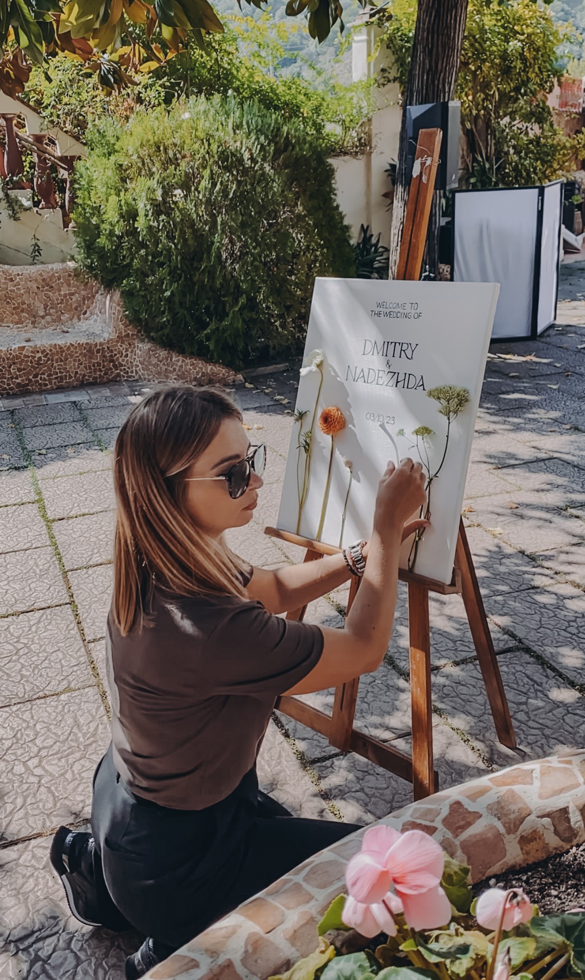 Girl with sunglasses painting on an easel outdoors, surrounded by trees and sunlight, with pink flowers in foreground.