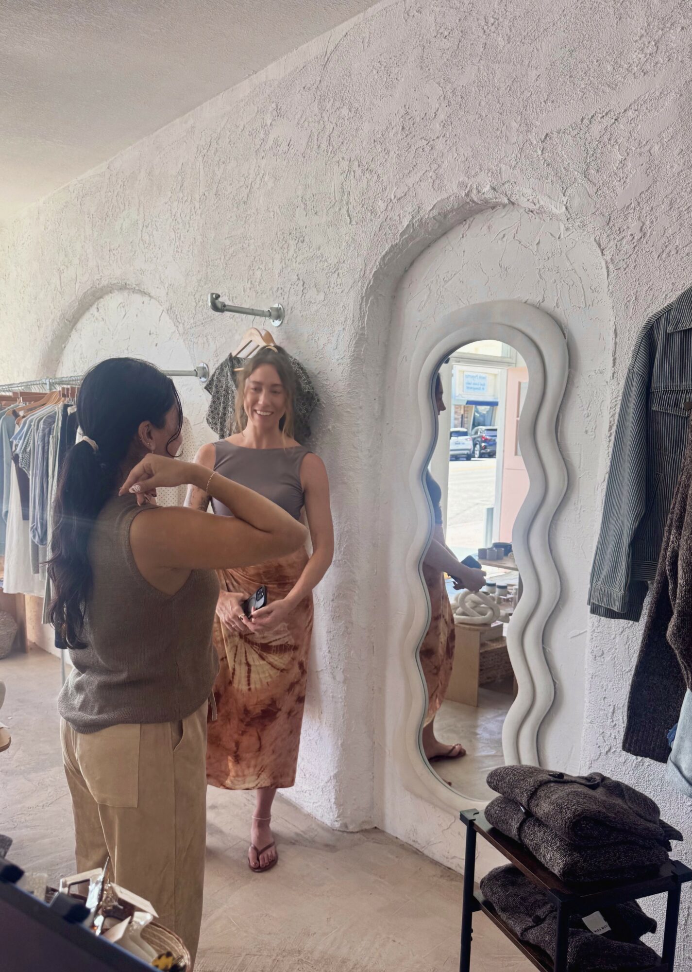 Two women in a clothing store, one adjusting the other's hair, with a mirror and clothing racks visible.