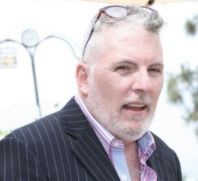 A middle-aged man with gray hair and beard, wearing glasses on his head, in a pinstripe suit and striped shirt, outdoors.