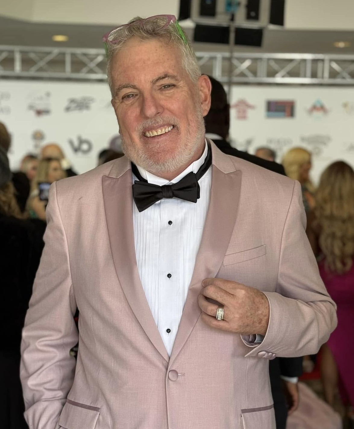 Man in tuxedo with pink blazer and black bow tie smiling at event, holding lapel, crowd in background.