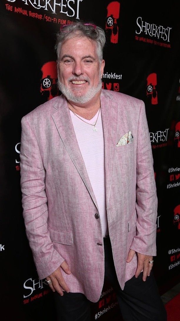 Smiling man with gray hair and beard in a pink blazer at an event, black background with red and white text