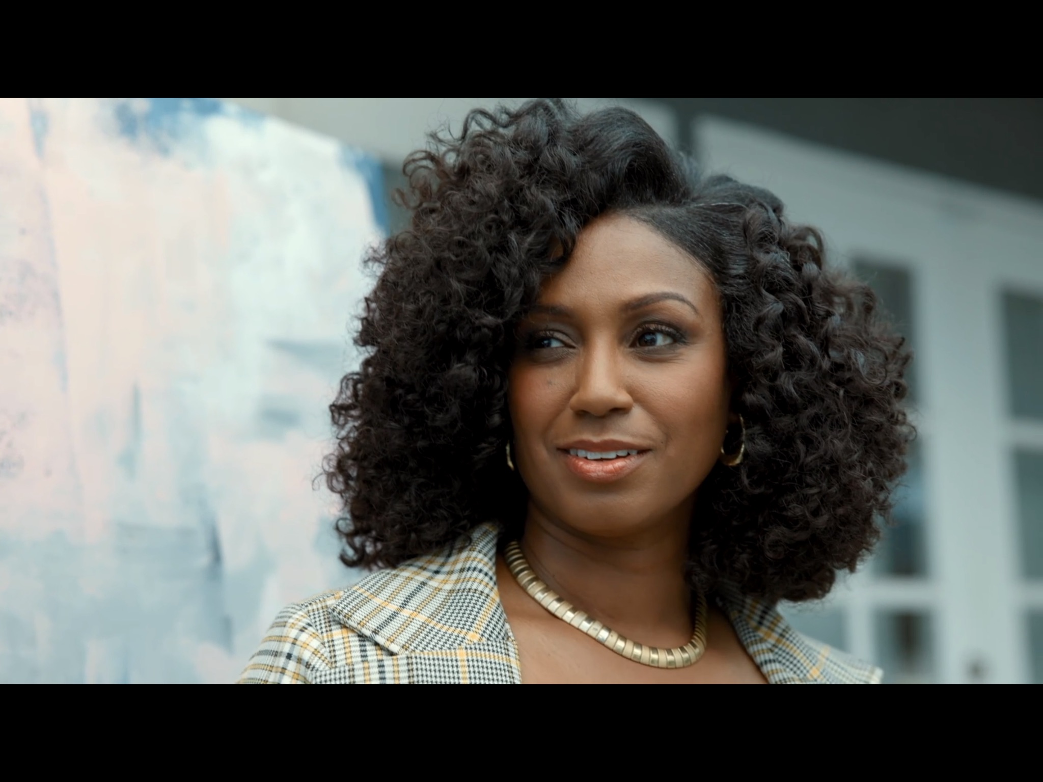 Woman with curly hair wearing a checkered blazer and pearl necklace, standing outdoors with a building in background.