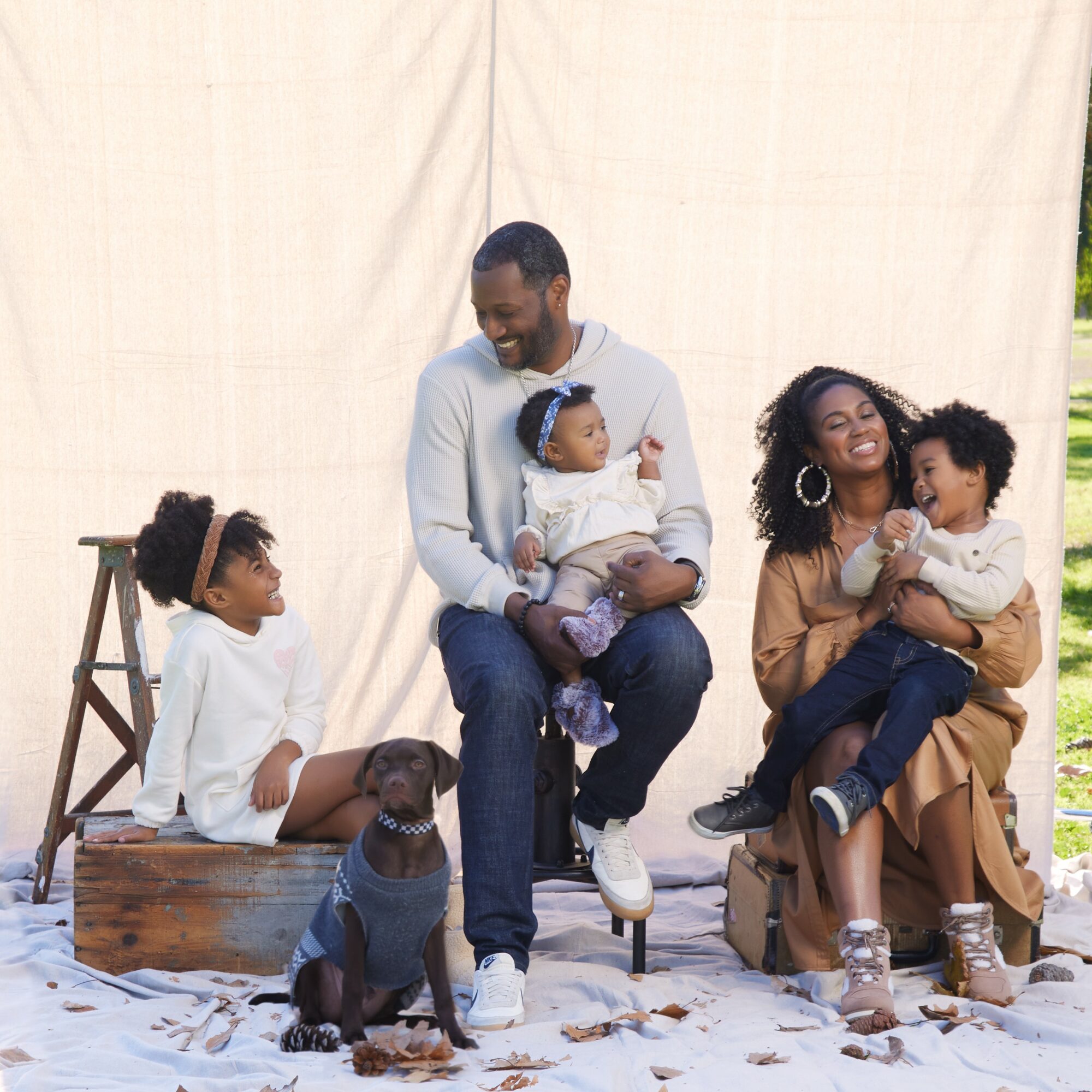 Family of four with two children and a dog sitting outdoors on a sandy surface, smiling and interacting happily.