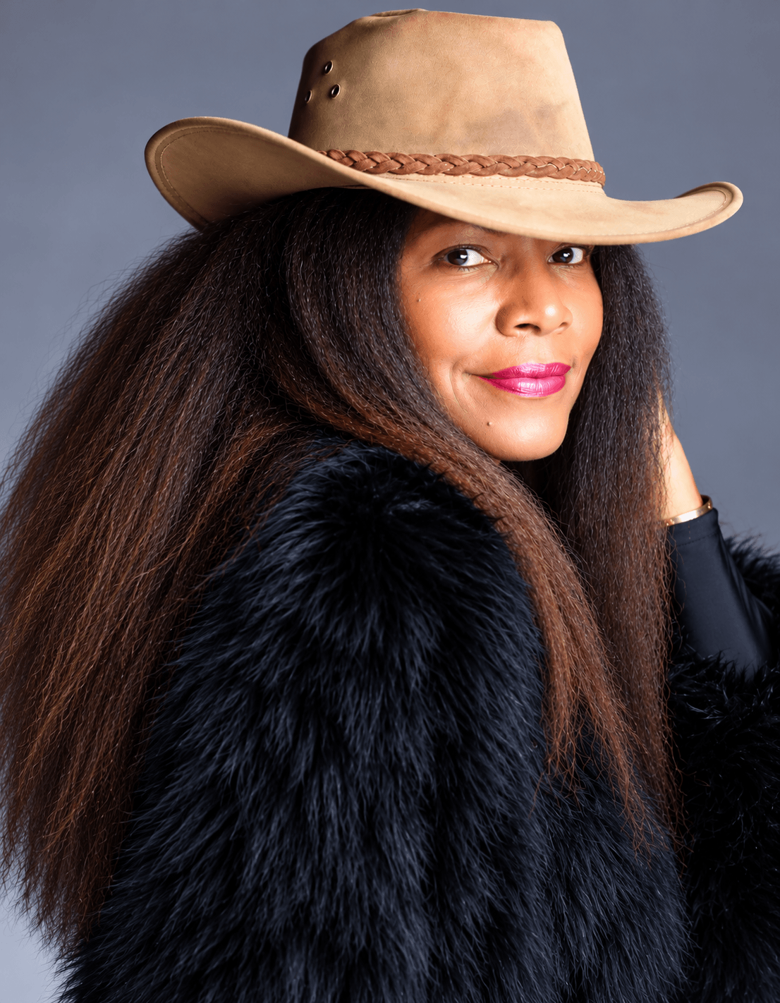 Woman wearing a beige cowboy hat and black fur coat, smiling, with long dark hair, against a gray background.