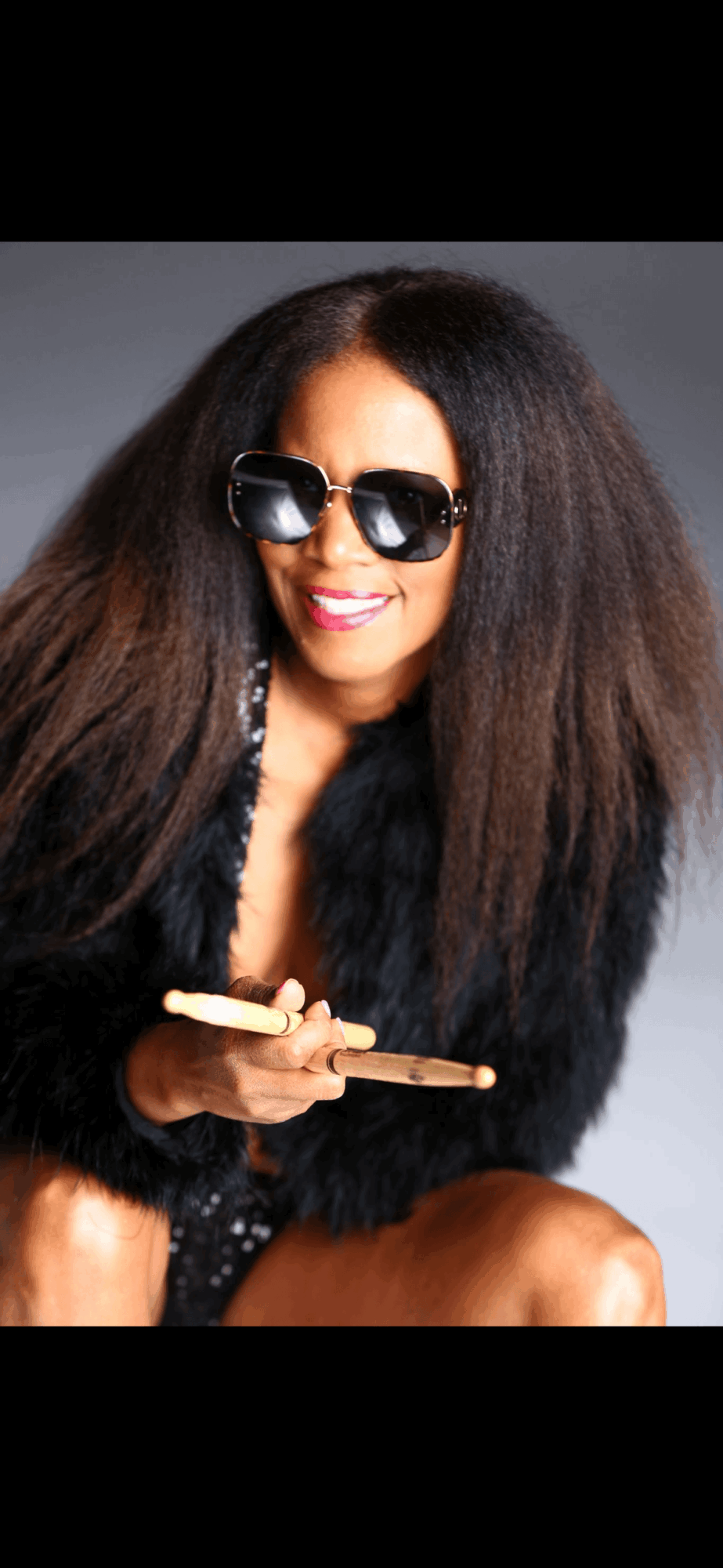 Woman with large sunglasses and voluminous hair smiling, holding a smartphone, against a plain background.