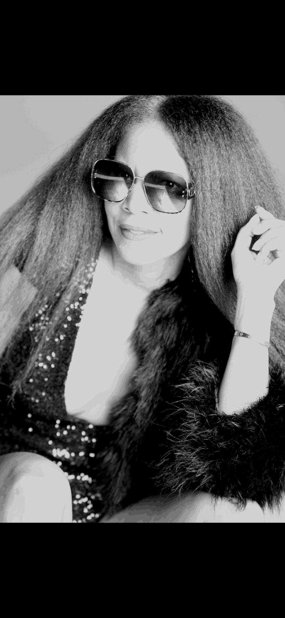 Woman with long hair wearing sunglasses and a sequined top, holding her hair, in black and white.
