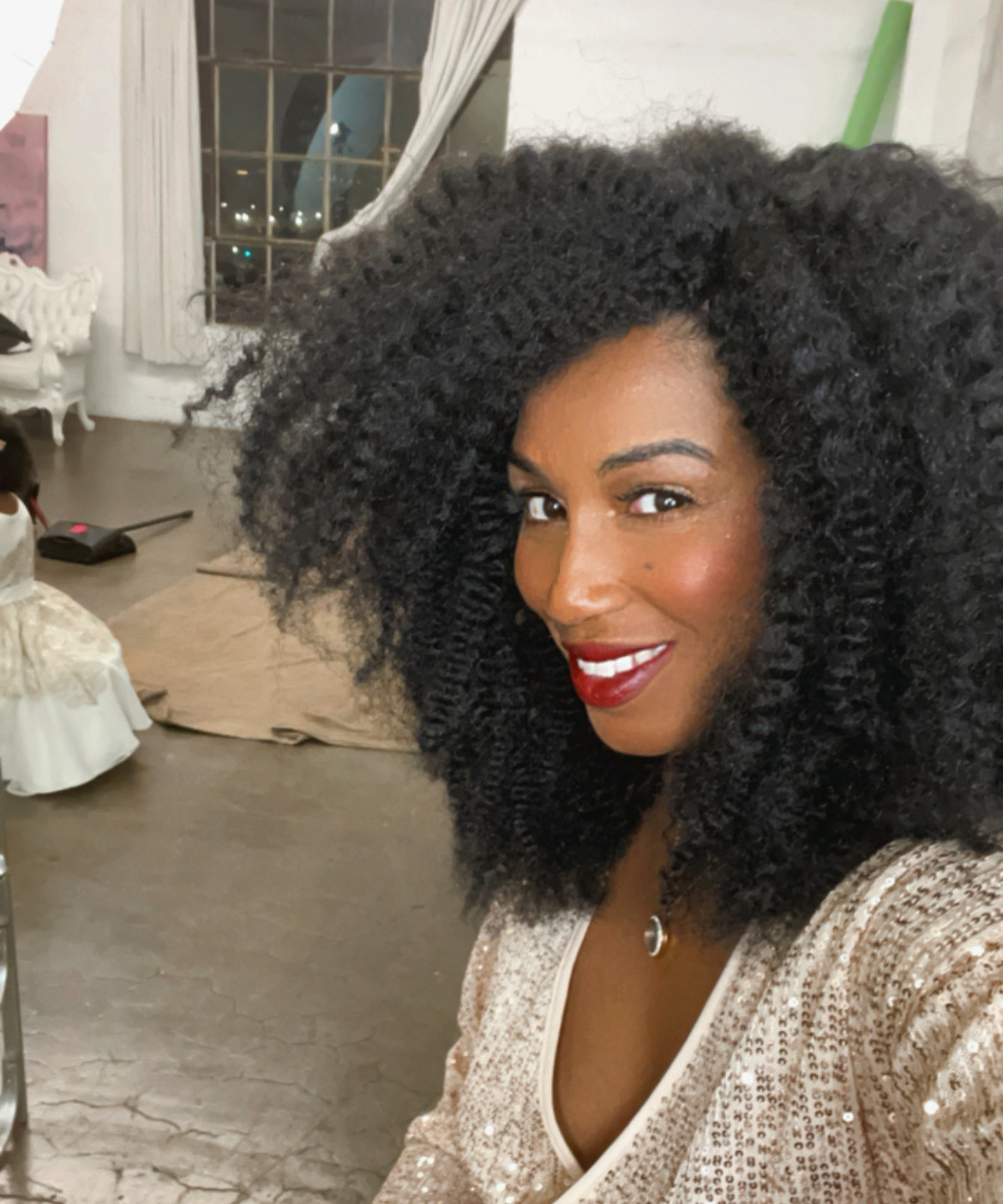 Woman with curly hair smiling indoors, wearing a light-colored top, in a room with a window and furniture.