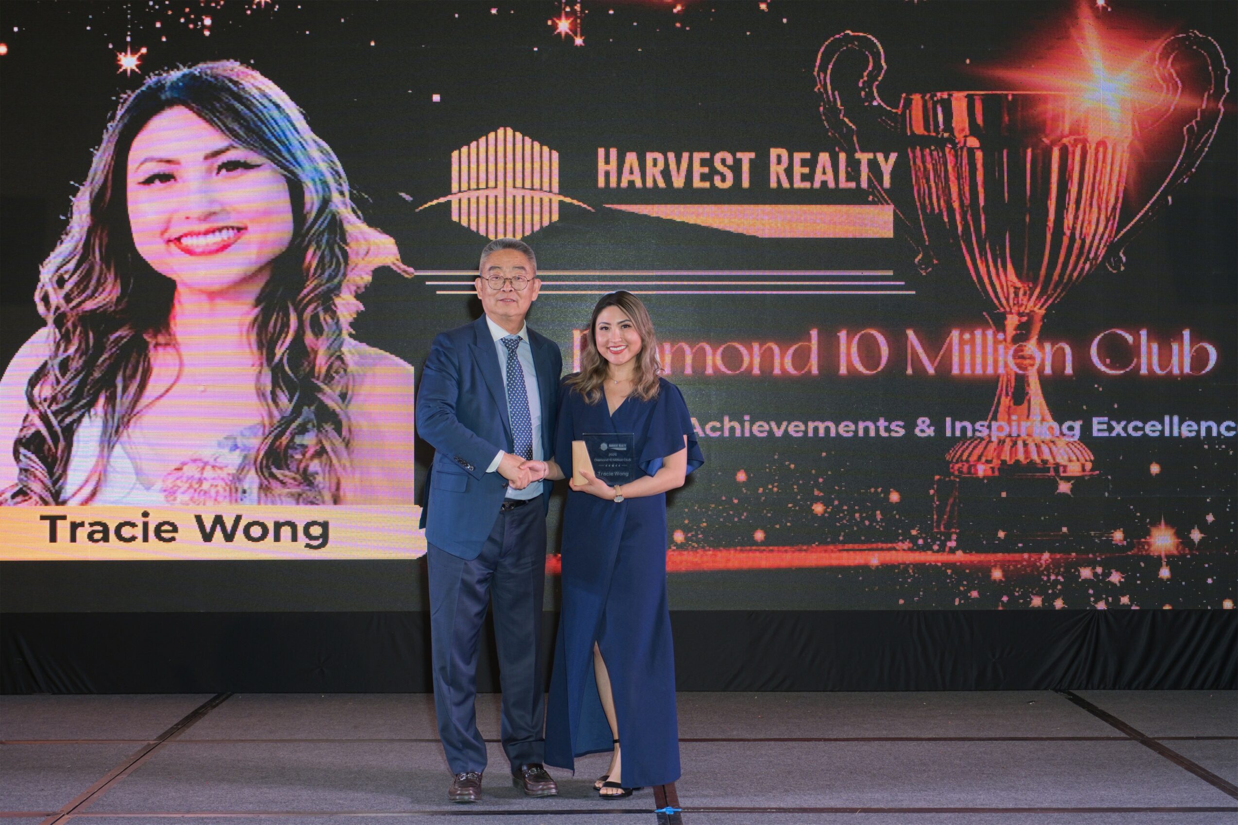Two people standing on stage in front of a large screen with a woman's photo, text, and a trophy graphic.