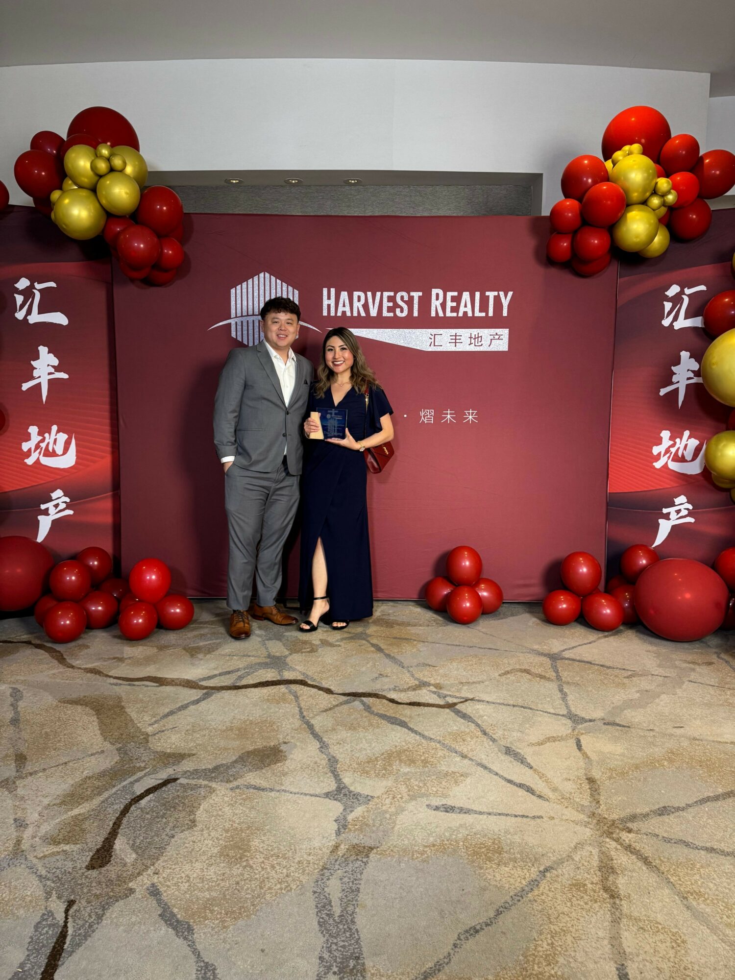 Two people standing in front of a red backdrop with decorative red and gold balloons, posing for a photo.