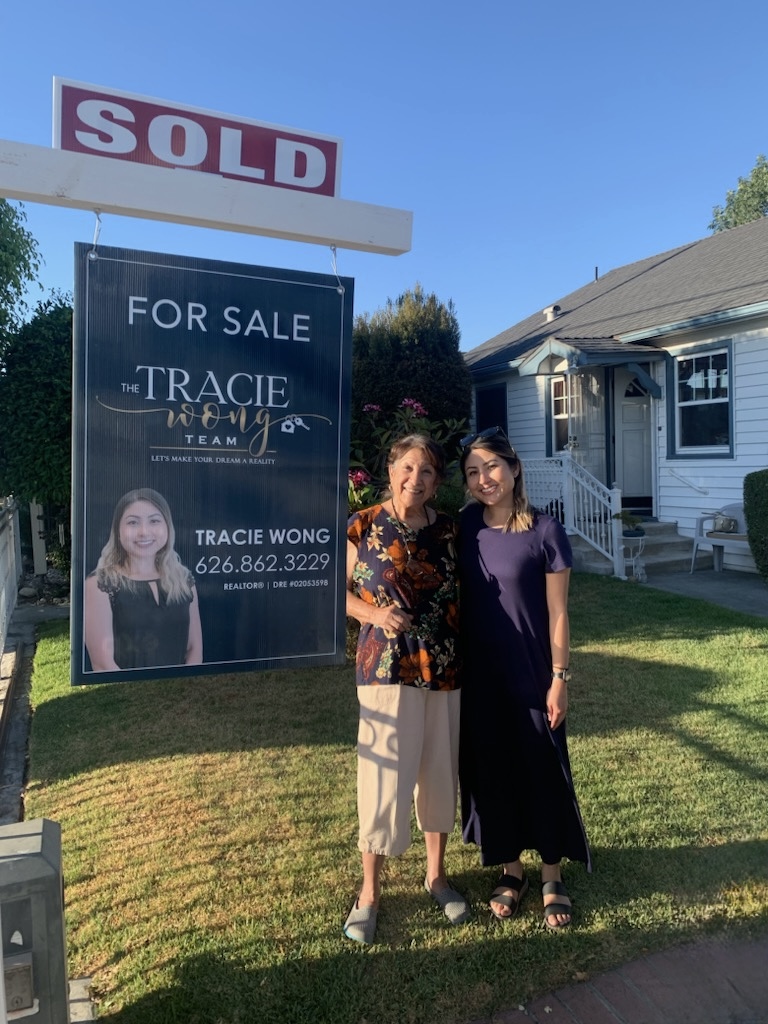 Two women stand in front of a real estate sign and a house, smiling outdoors on a sunny day.