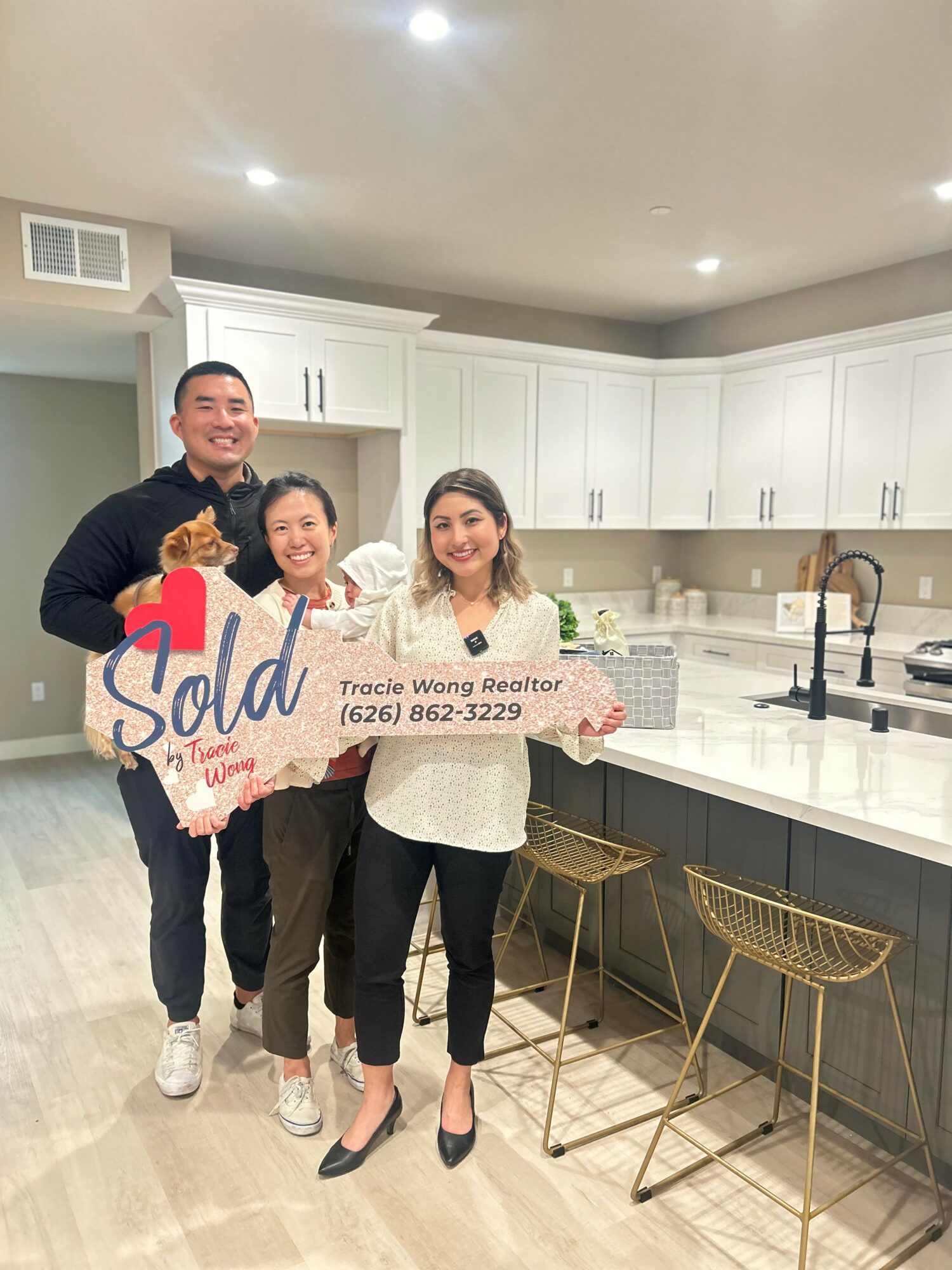 Three people standing in a kitchen holding a 'Sold' sign, smiling, with white cabinets and a countertop in the background.