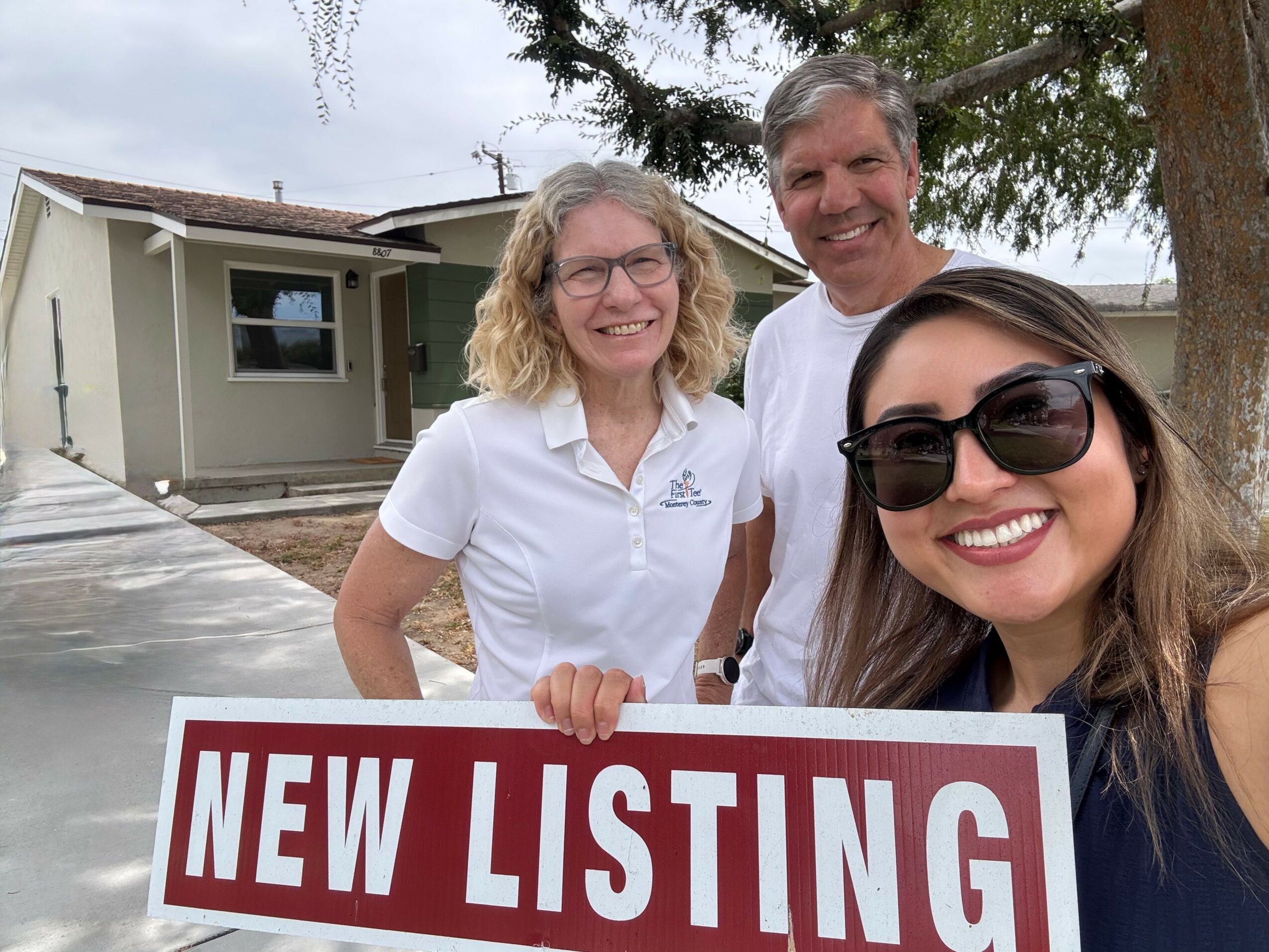 Three people smiling outdoors holding a sign that says 'NEW LISTING' in front of a house with trees and a cloudy sky.