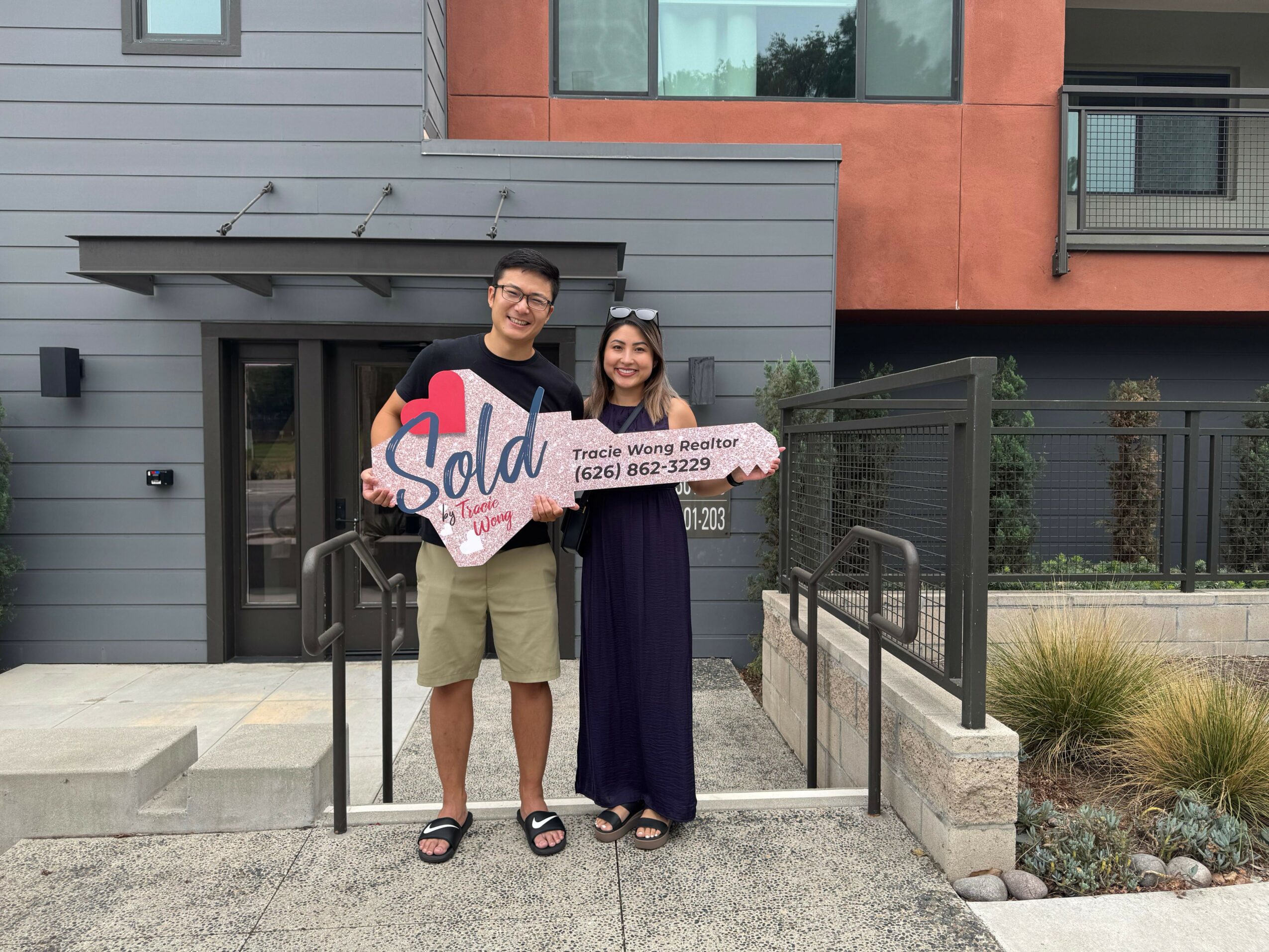Two people holding a pink and white sign with the word 'SOLD' in front of a house, smiling, with a woman in a purple dress.