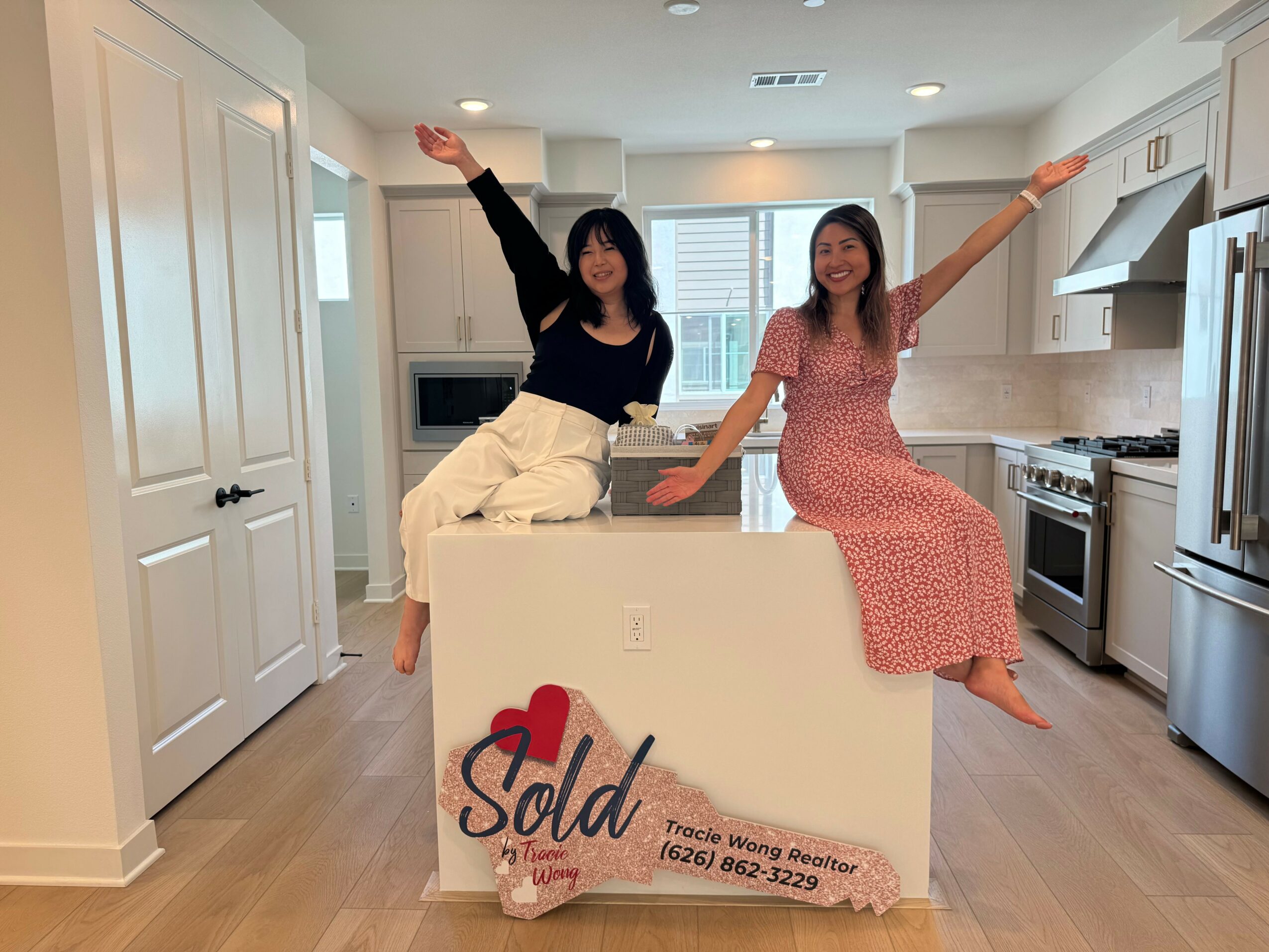 Two women sitting on a large white sign in a kitchen, smiling and raising their arms, with a sold sign in front.