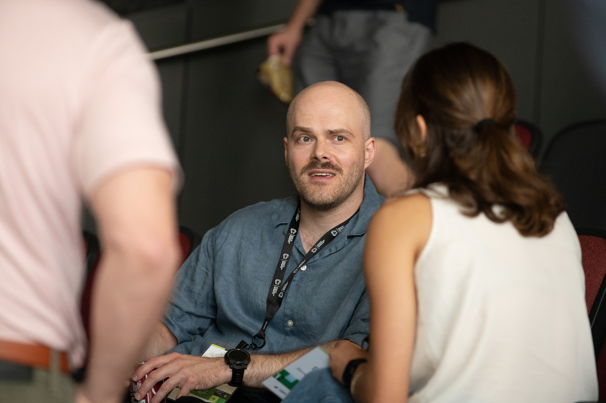 Bald man with a beard wearing a denim shirt, talking to a woman with brown hair in a white top, in a social setting.