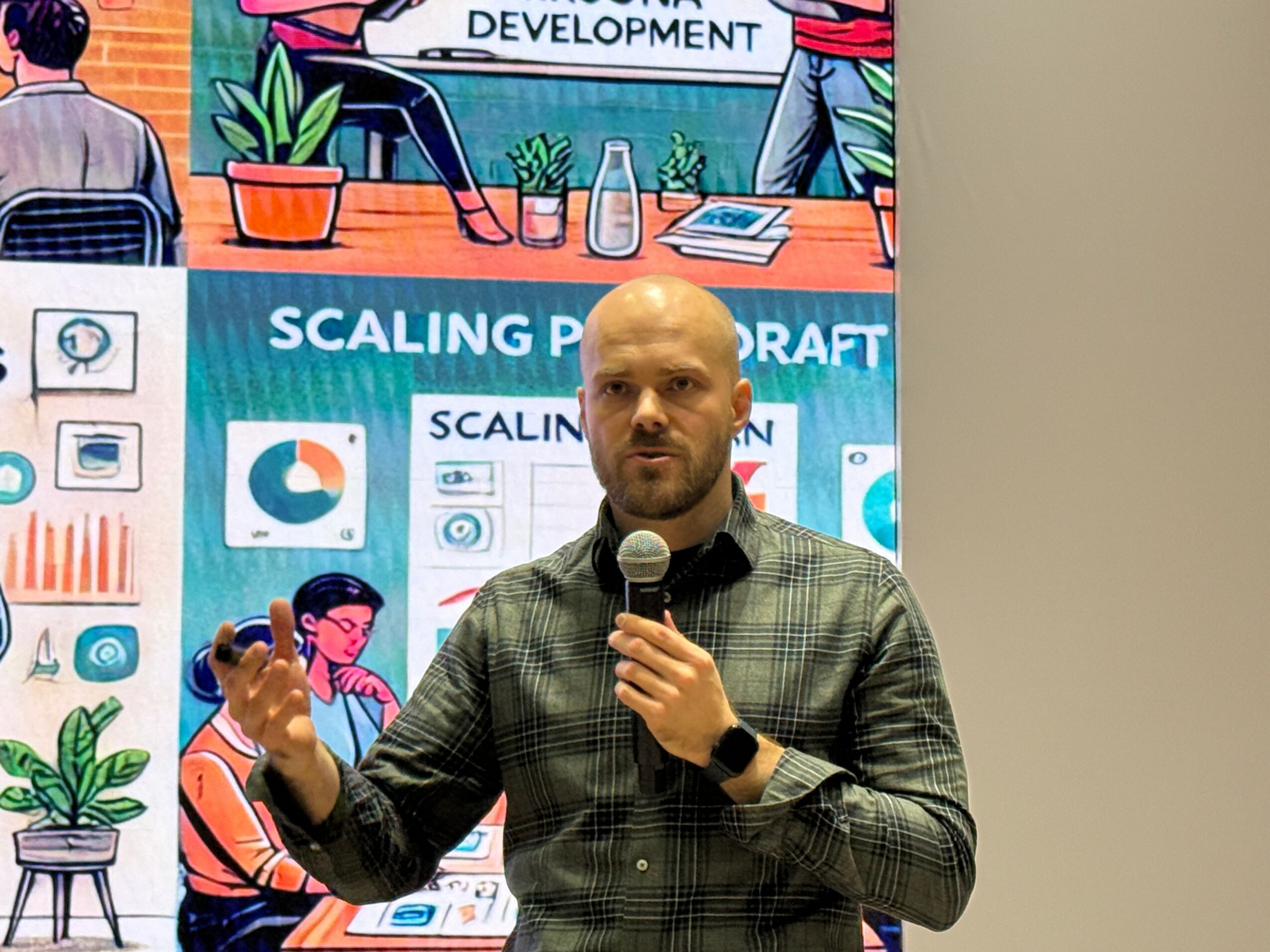 Man with a beard and bald head speaking into a microphone, standing in front of a presentation screen, gesturing with his left hand.