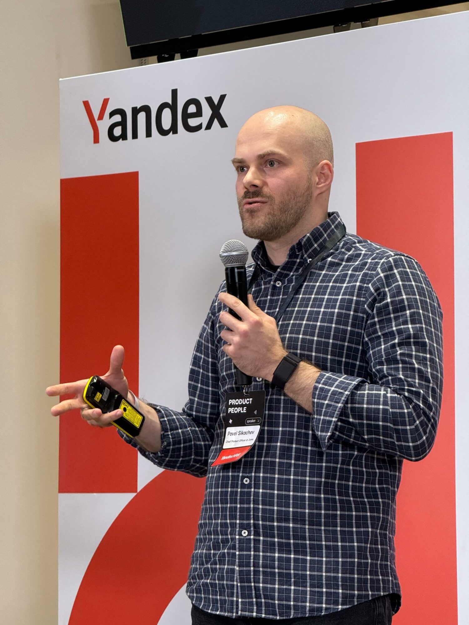 Man with a shaved head wearing a checkered shirt, holding a microphone and a remote, standing in front of a Yandex banner.