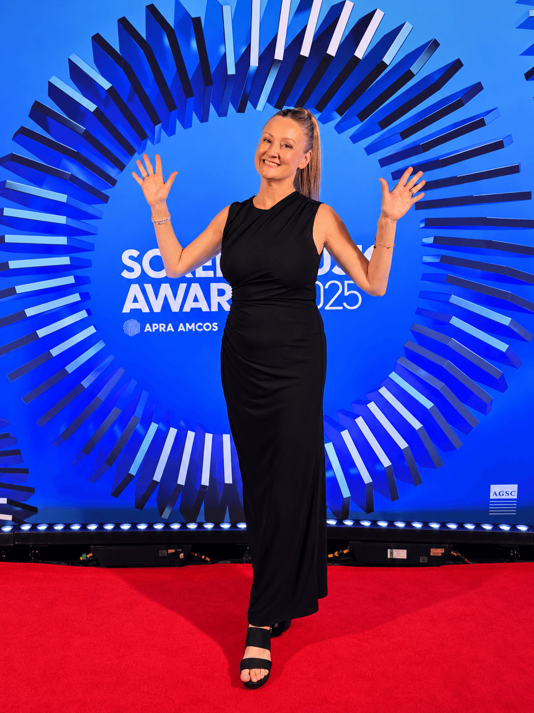 Woman in black dress standing on red carpet, smiling, with blue circular backdrop behind her, at an awards event.