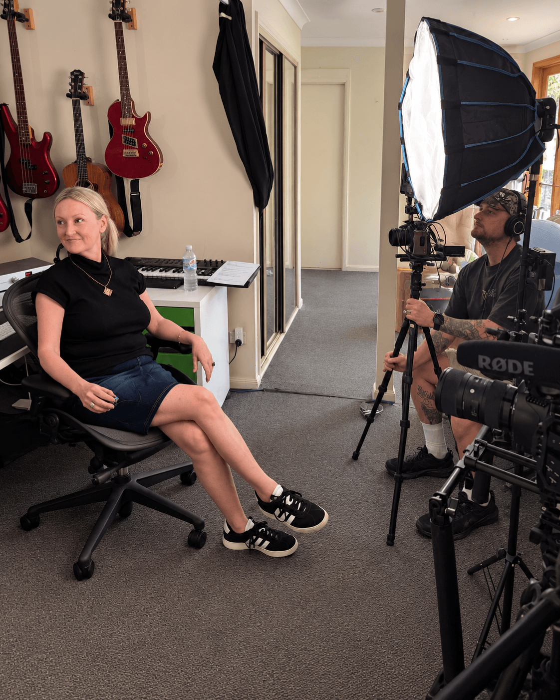 Woman sitting in a chair during a video shoot, with guitars hanging on the wall behind her, and filming equipment around her.