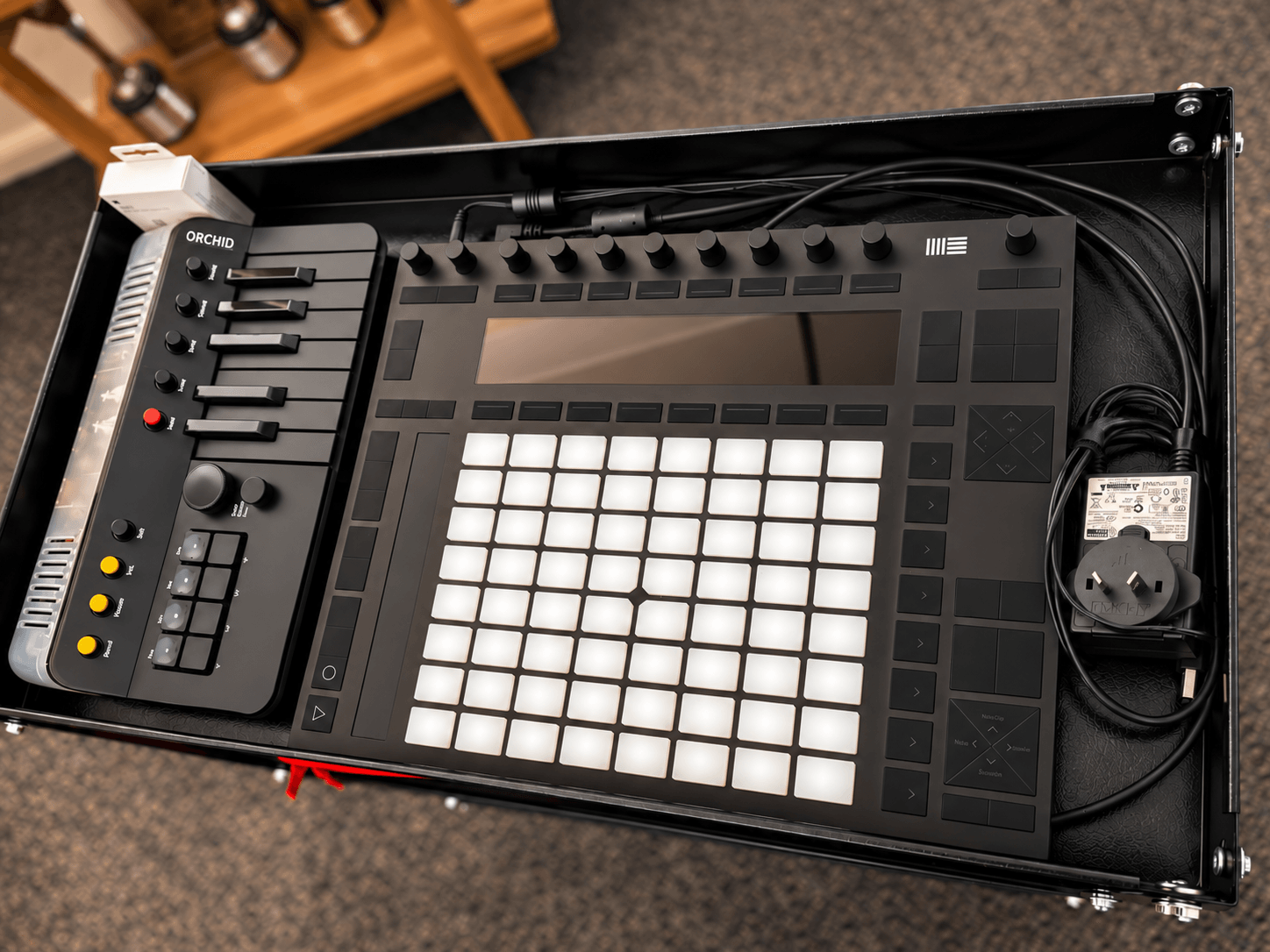 Music production equipment with a grid controller, mixer, and cables on a black surface.